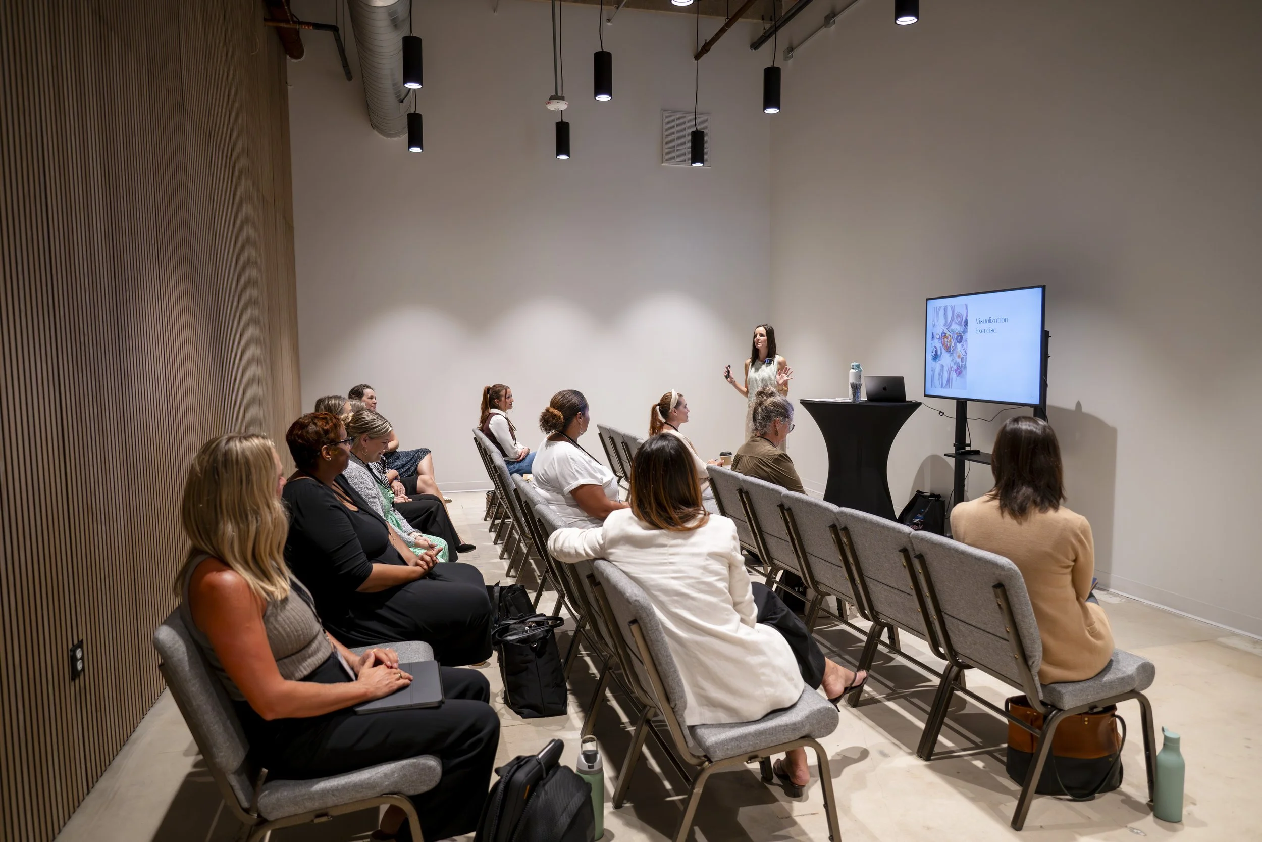 A woman giving a presentation to a group of people in a conference room with white walls and a wooden accent wall. The audience is seated in gray chairs, and a large screen displays a slide with some text and images.