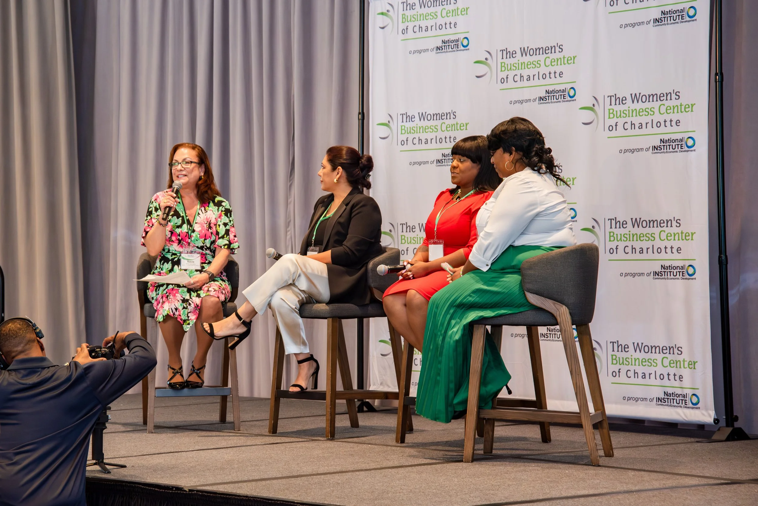 Four women participating in a panel discussion at a conference, with one woman speaking into a microphone and the others listening, on a stage with a backdrop displaying logos for The Women's Business Center of Charlotte and the National Institute.