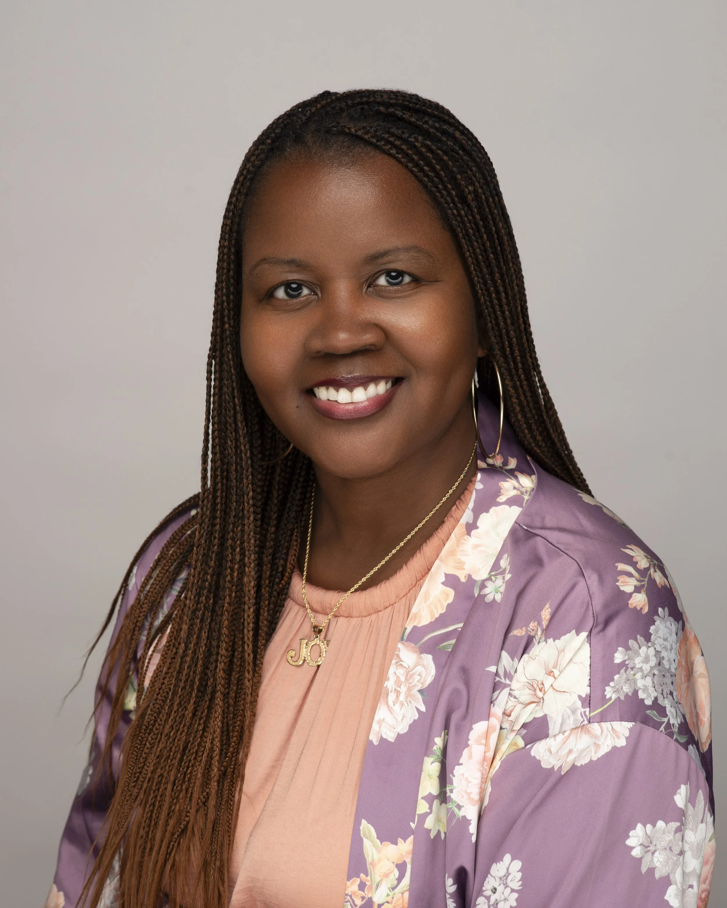 A smiling woman with long braided hair, wearing a pink top, a purple floral jacket, gold hoop earrings, and a gold necklace with a 'Joy' pendant, against a plain light gray background.