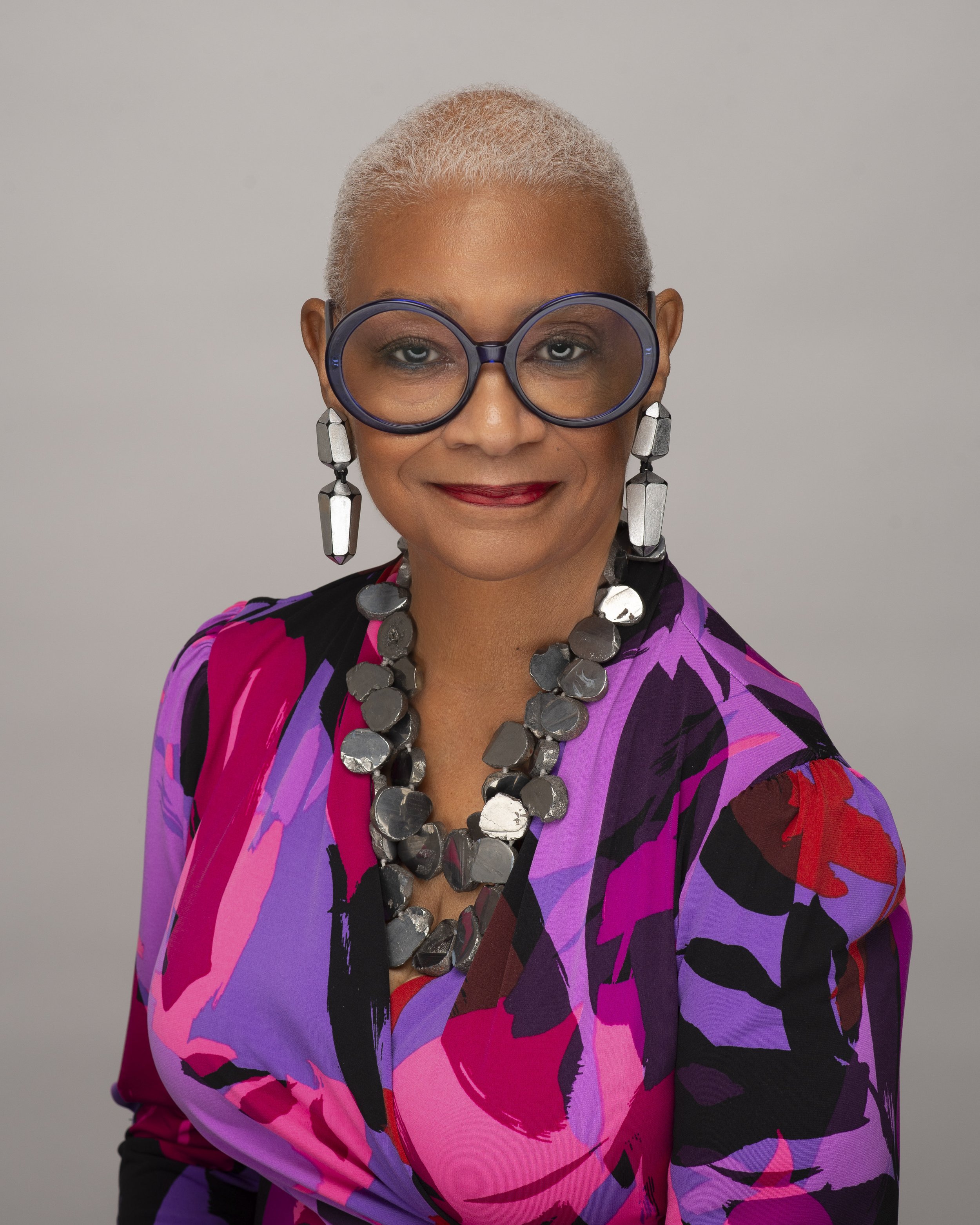 Portrait of a woman with short, platinum blonde hair, wearing large blue glasses, bold earrings, a chunky necklace, and a colorful purple, pink, and black patterned blouse, against a neutral background.