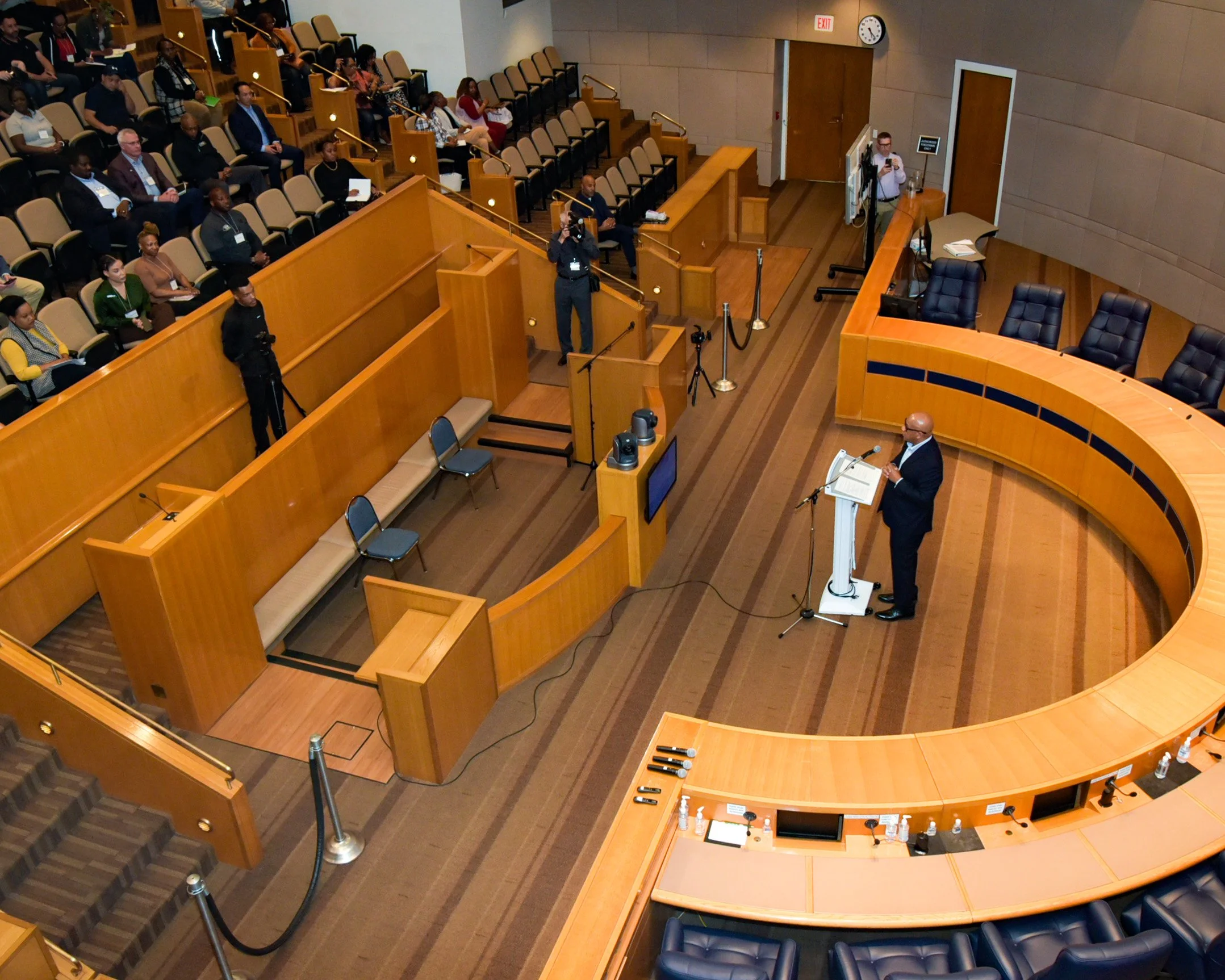 A man in a suit and glasses speaking at a podium in a large conference room with curved wooden paneling, an audience seated in rows, and photographers taking pictures.