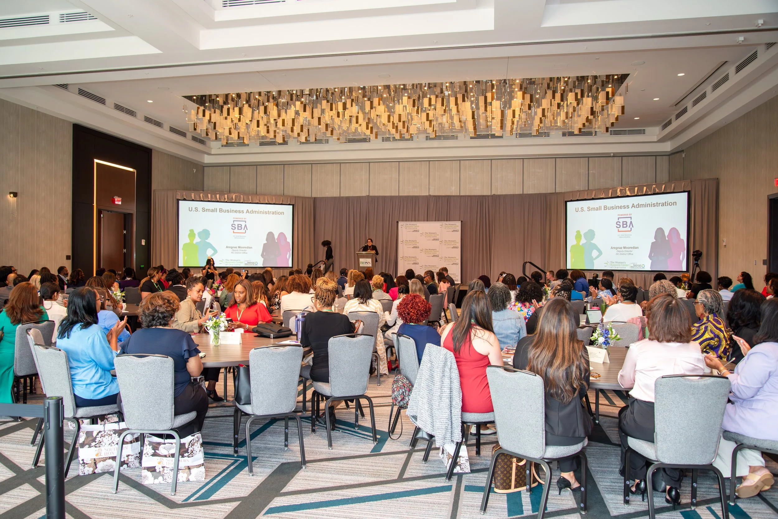 A conference room filled with women attending a presentation on U.S. Small Business Administration, with two large screens displaying the presentation slides, flowers on the tables, and a speaker at the podium.