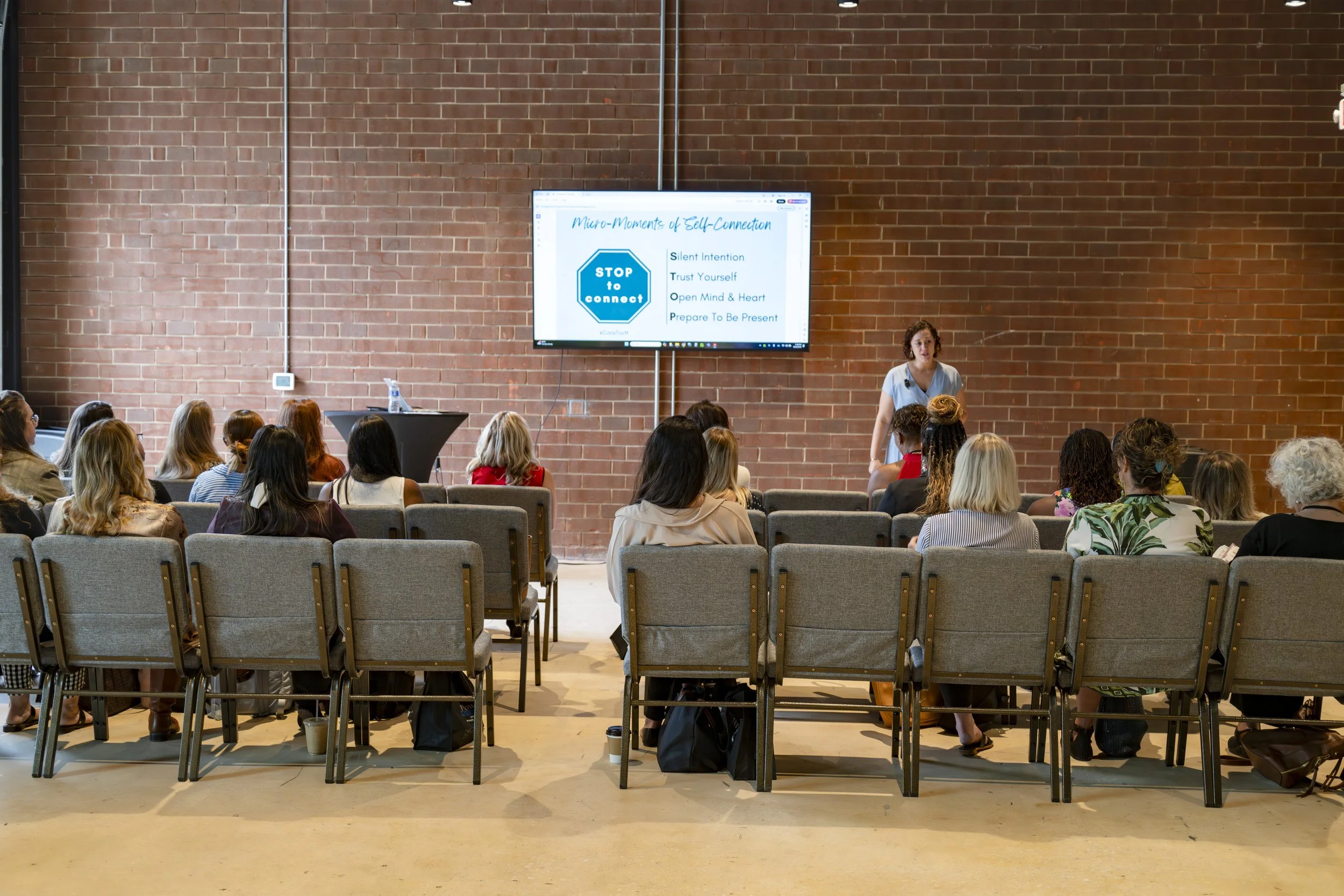 A woman giving a presentation to an audience in a room with a brick wall, standing in front of a large screen displaying a slide titled 'Micro-Moments of Self-Connection.' The slide lists four points: Silent Intention, Trust Yourself, Open Mind & Hea