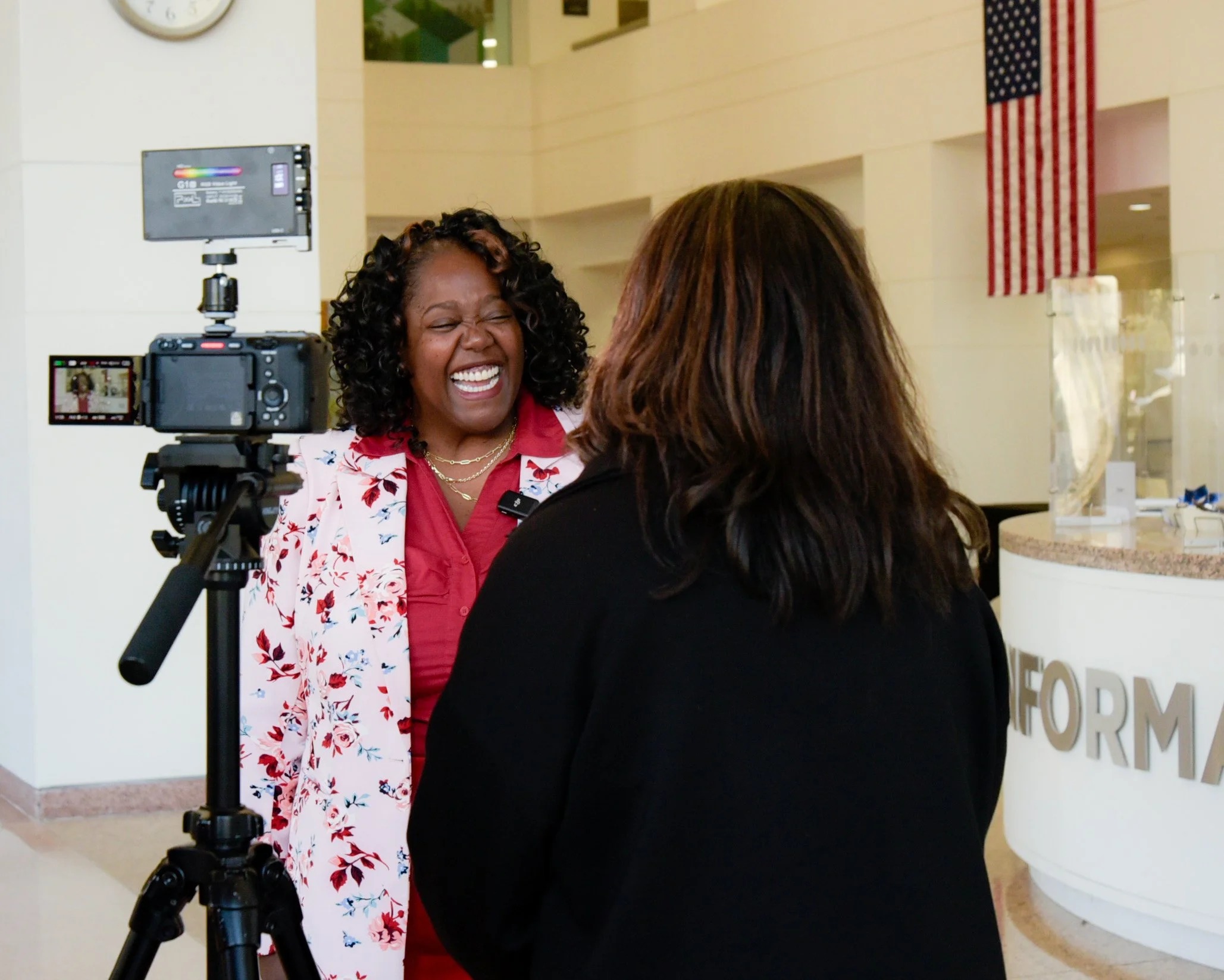 A woman with curly hair wearing a floral blazer laughs while being interviewed by another woman with brown hair at a registration or information desk in a public building. A camera on a tripod is set up to record the interview.