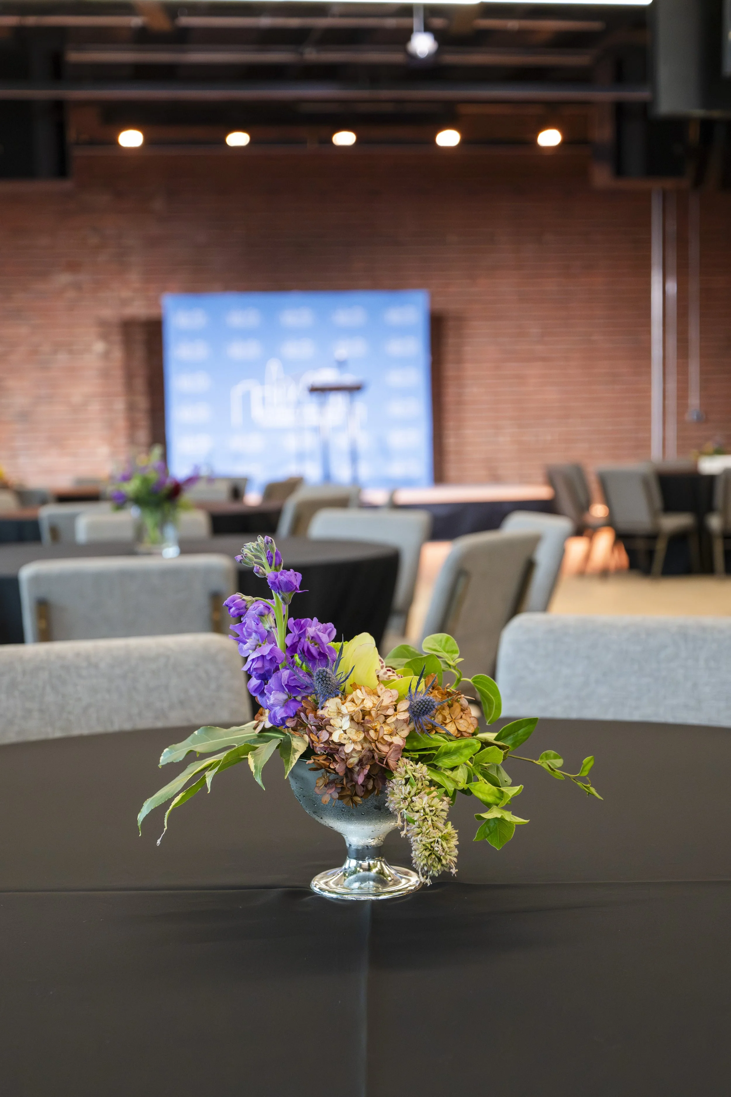 A floral centerpiece with purple and green flowers on a black table in a conference room or event space with a stage and chairs in the background.