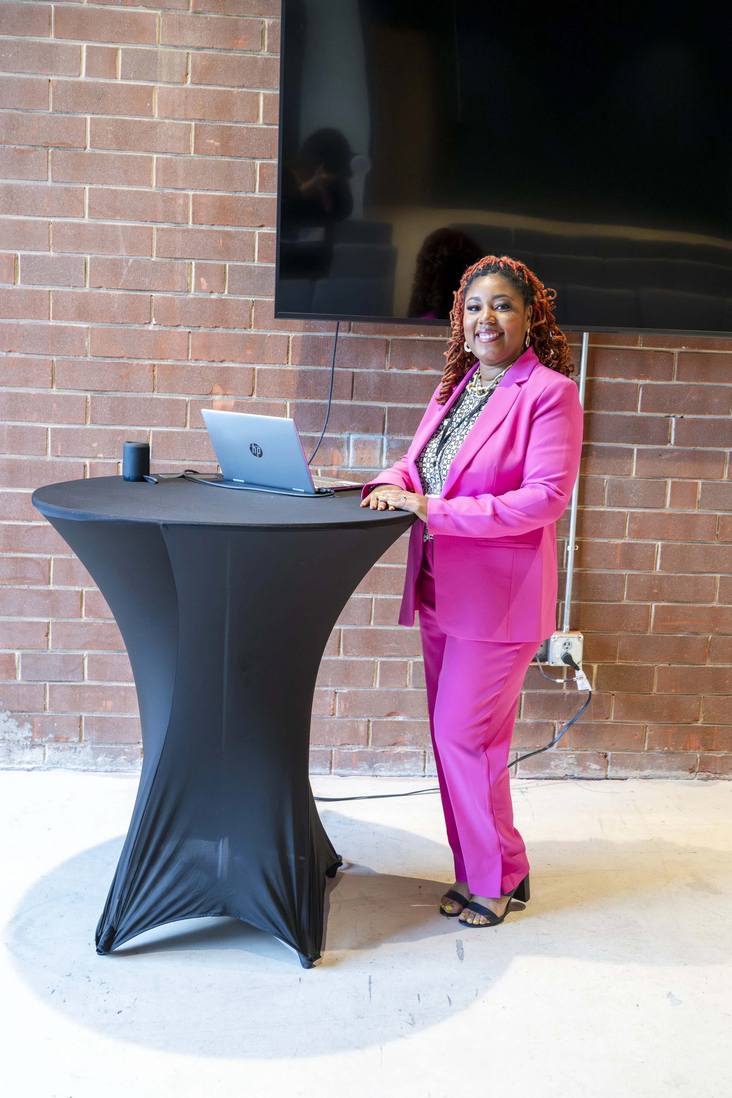 A woman in a bright pink suit smiling while standing behind a black covered high table with a laptop on it, in front of a brick wall and a large television screen.