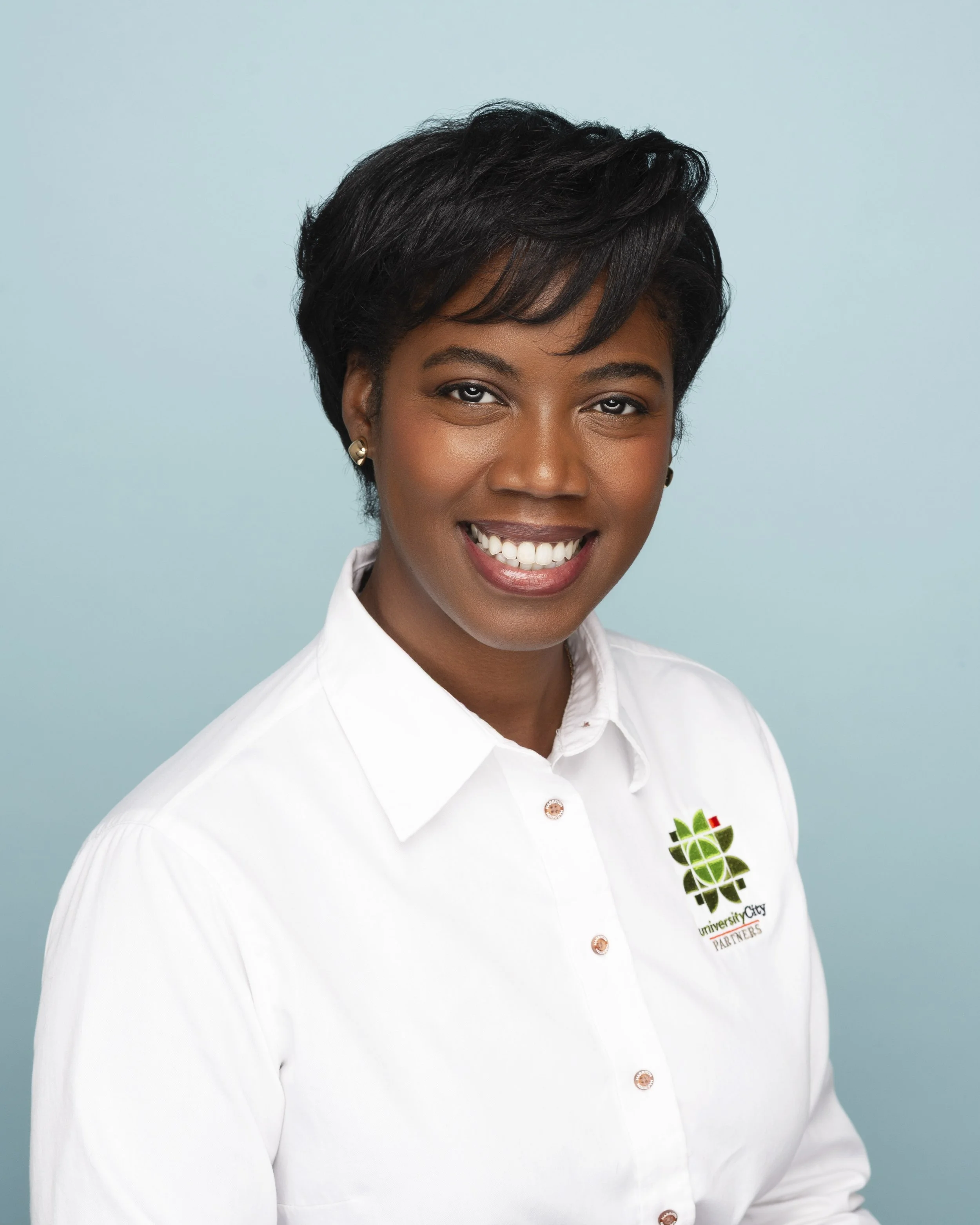 A woman with short black hair smiling, wearing a white button-up shirt with a logo on the left side that reads 'University City Partners'.
