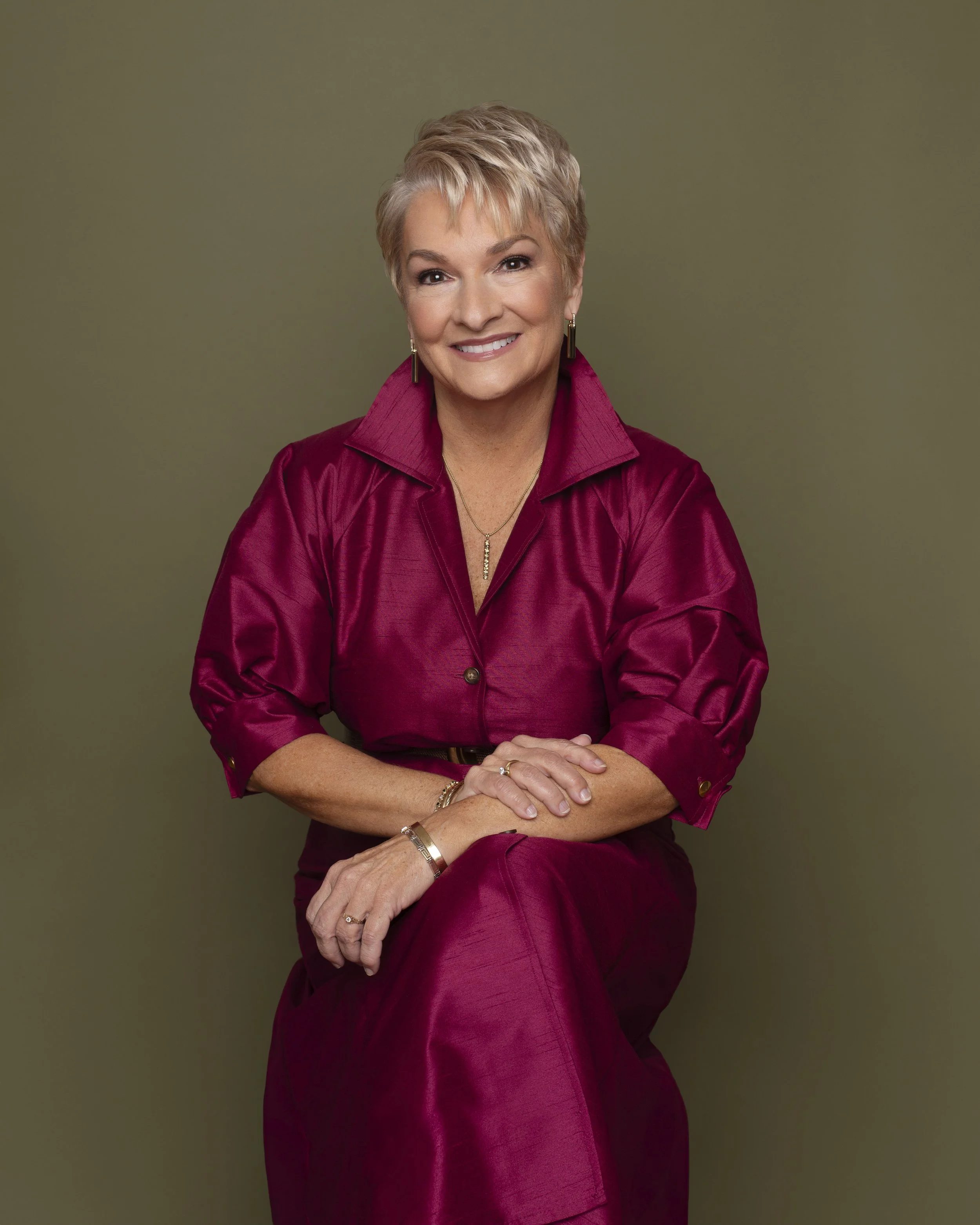 An older woman with short, gray hair, smiling, wearing a maroon silk blouse and matching skirt, sitting against a plain olive green background.