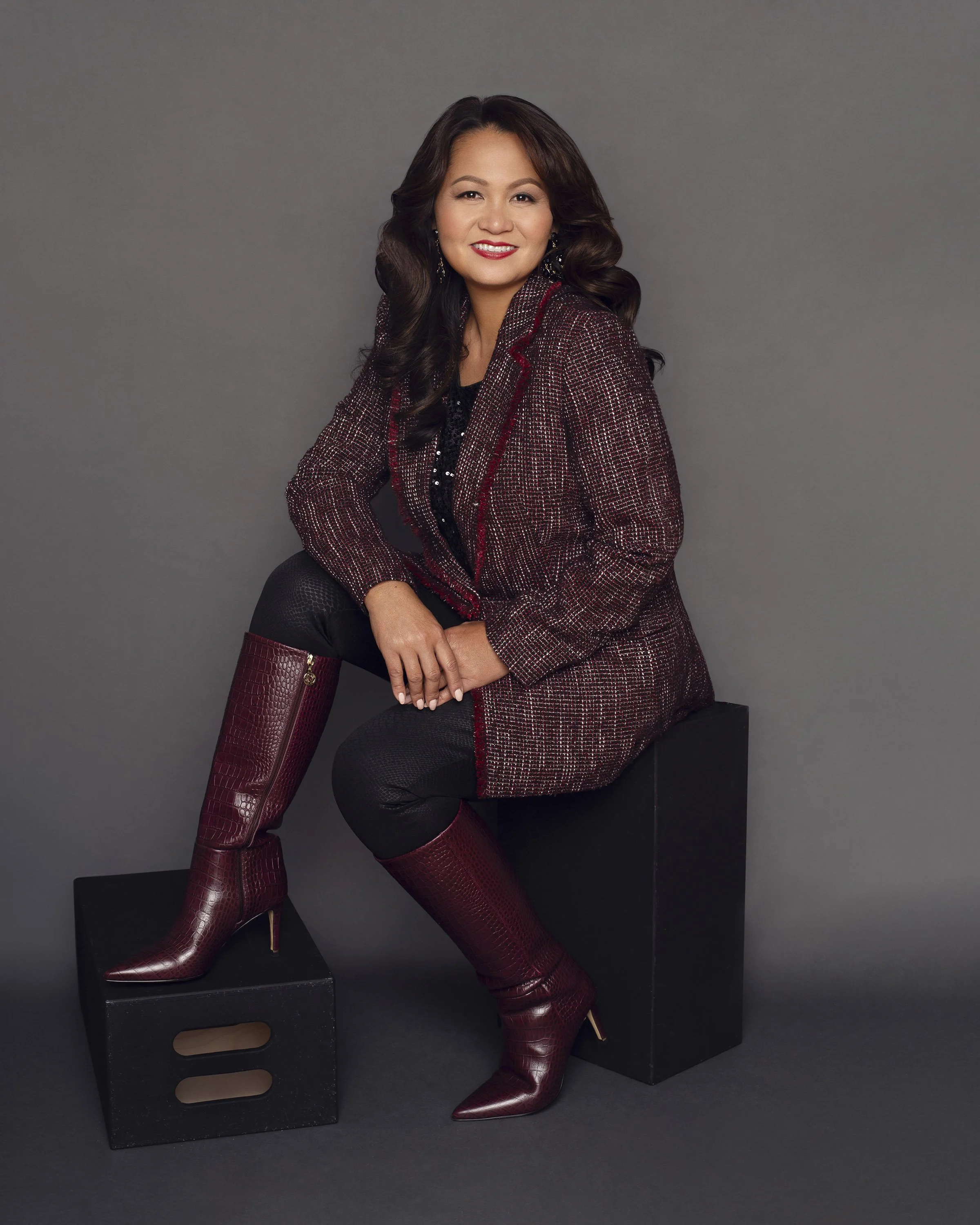 A woman with long wavy hair wearing a burgundy and black blazer, black pants, and matching high-heeled boots, sitting on black blocks against a gray background.