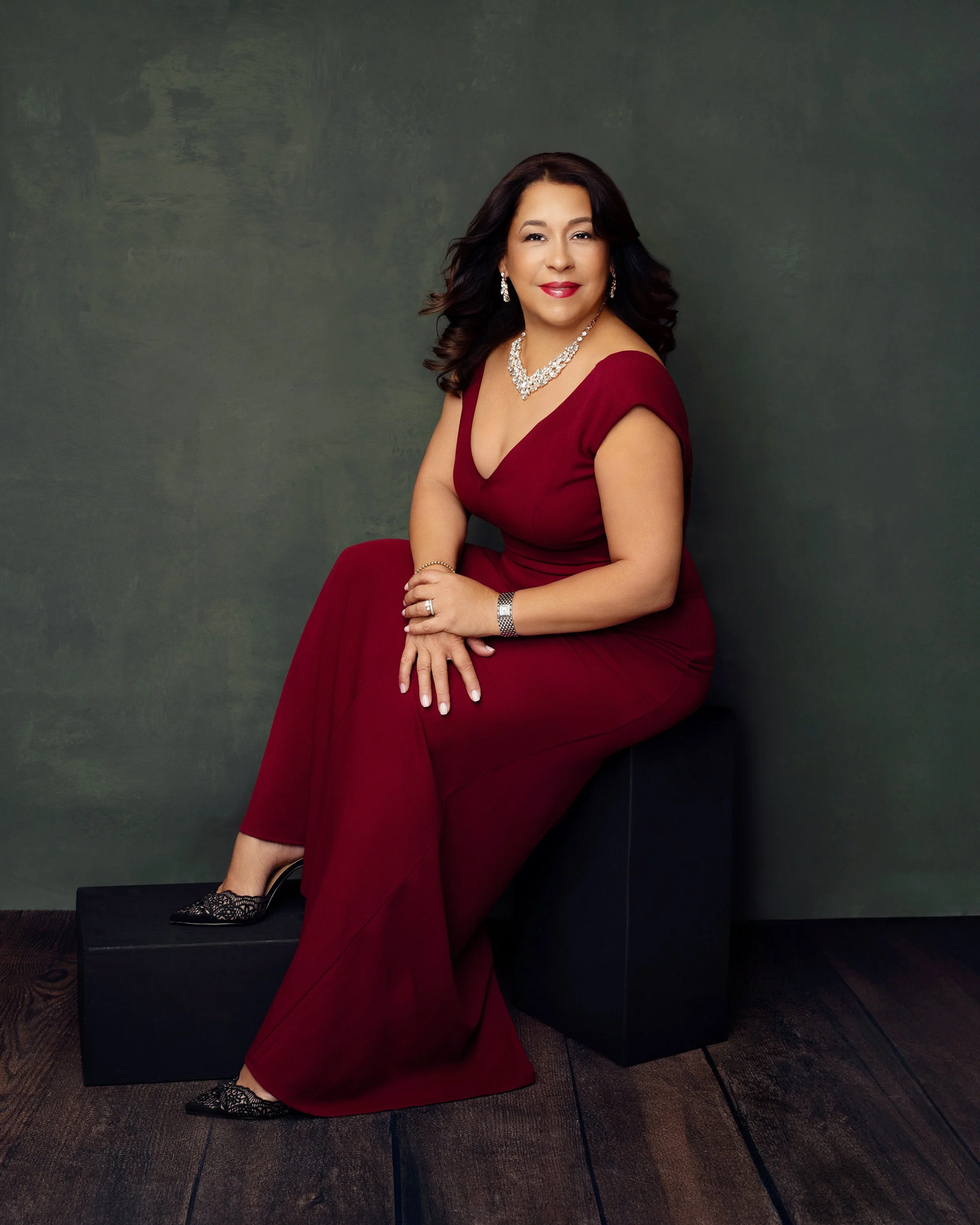 A woman in a red evening gown sitting on a black block against a grey-green background, wearing jewelry and black lace shoes