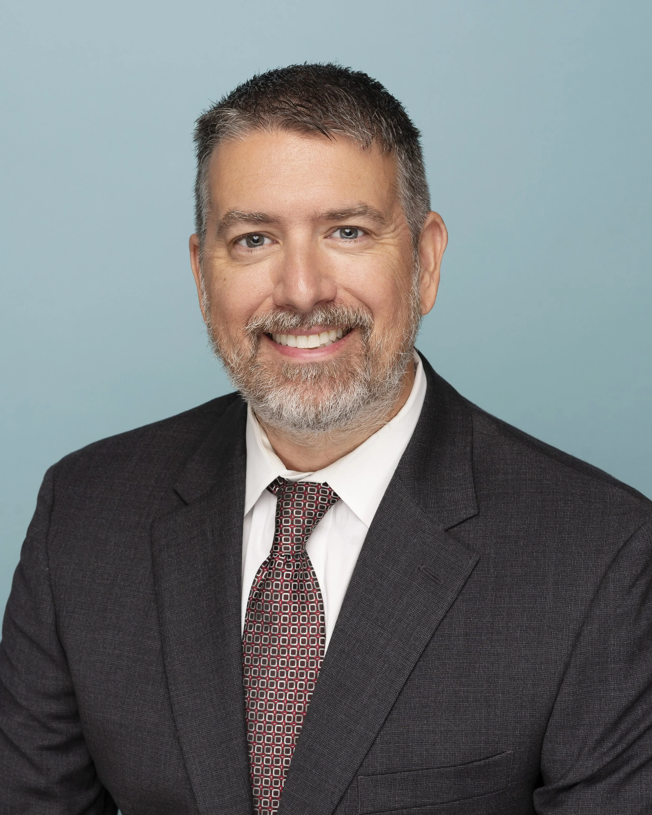 Headshot of a smiling middle-aged man with gray hair and beard, wearing a dark suit, white shirt, and patterned tie, against a light blue background.