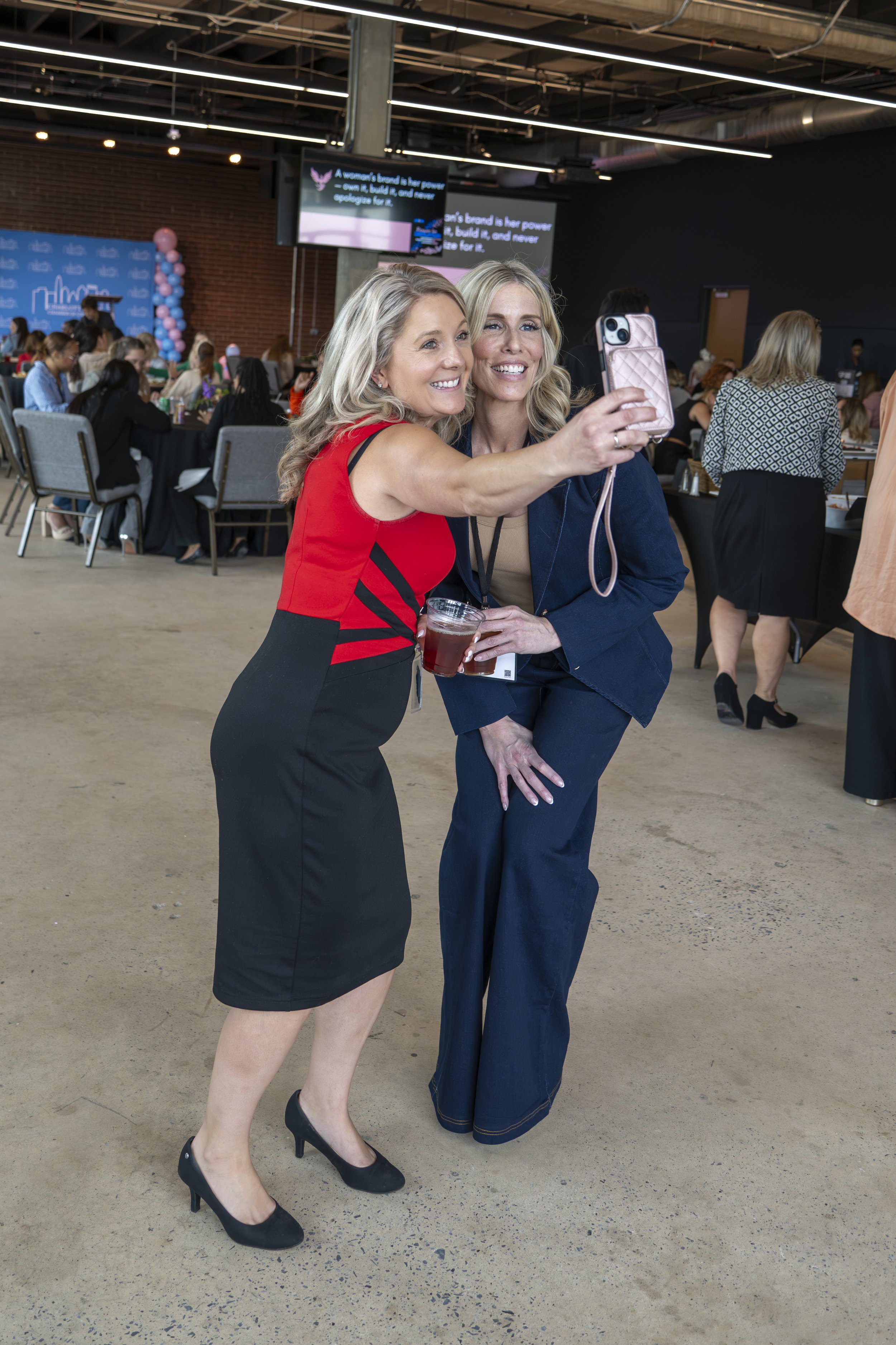 Two women taking a selfie at a professional event or conference, with a crowd of seated attendees in the background.