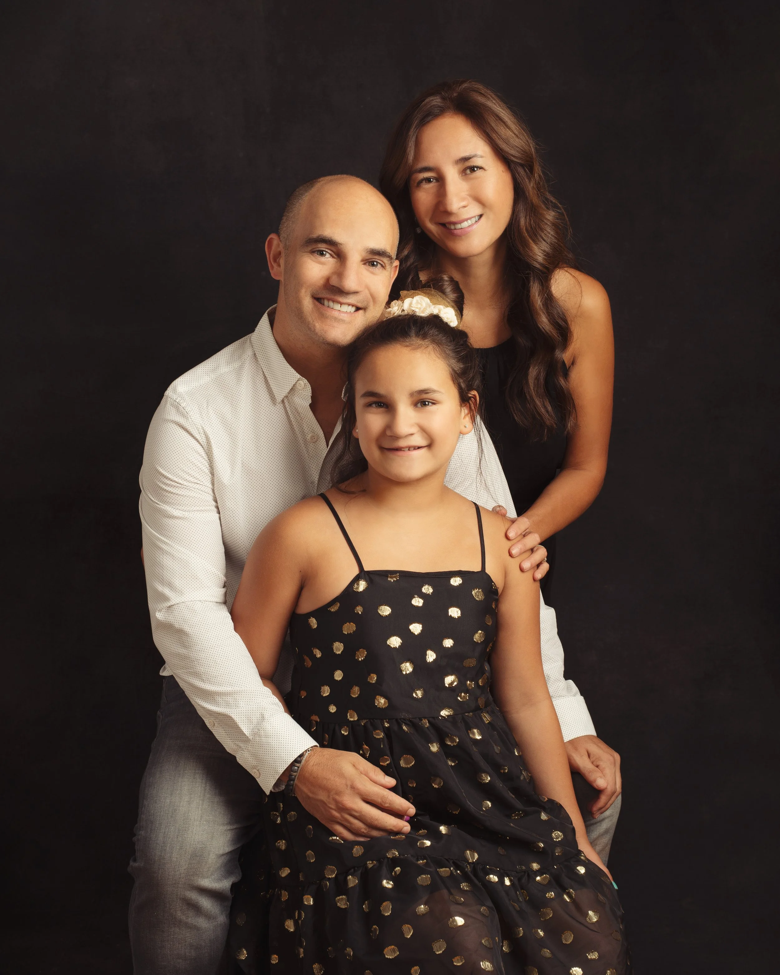 A family of four posed together in front of a dark background. The father is seated, wearing a white shirt, with the mother standing behind him. A young girl in a black dress with gold polka dots sits on his lap. The family is smiling.
