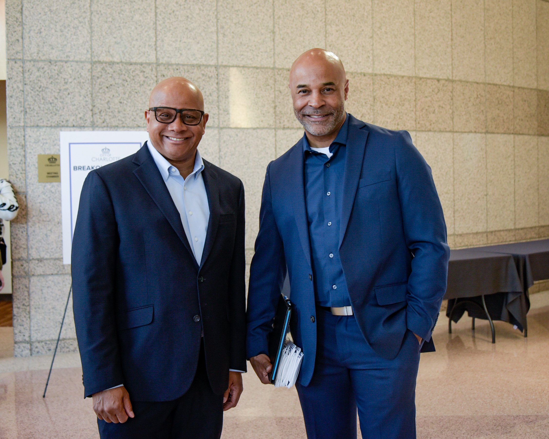 Two men in blue suits smiling and standing in an indoor setting. One man holds a notebook.