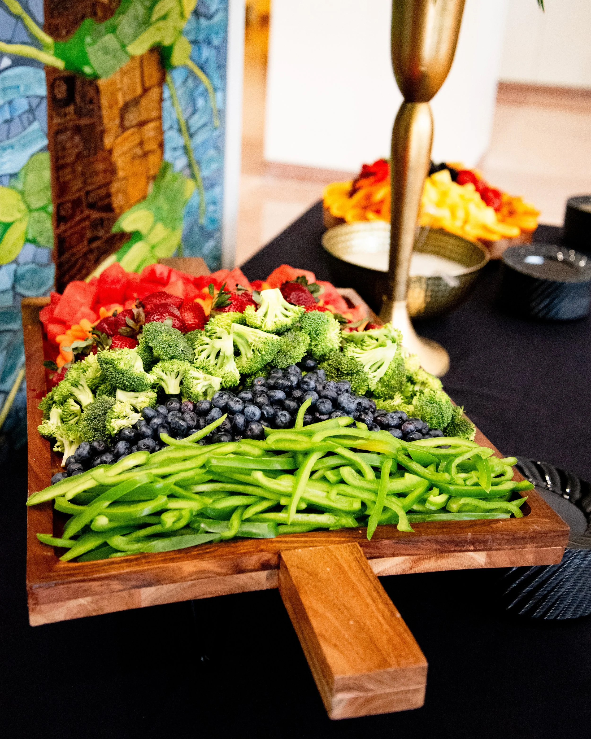 Wooden serving tray with fresh broccoli, blueberries, green beans, strawberries, and watermelon slices. Background includes bowls of sliced vegetables and decorative items on a black table.