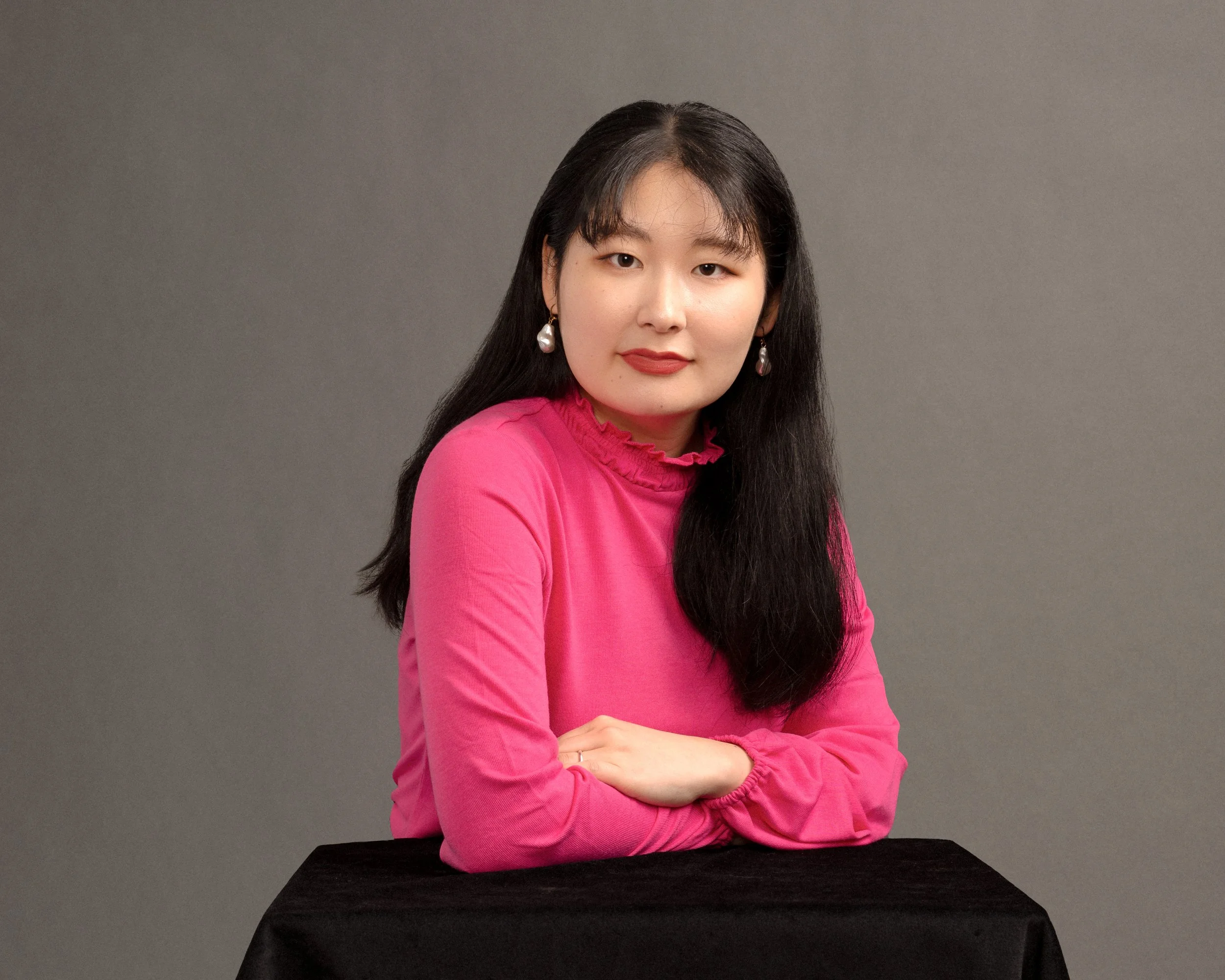 Portrait of a young Asian woman with long black hair, wearing a pink top and pearl earrings, sitting at a black table against a gray background.
