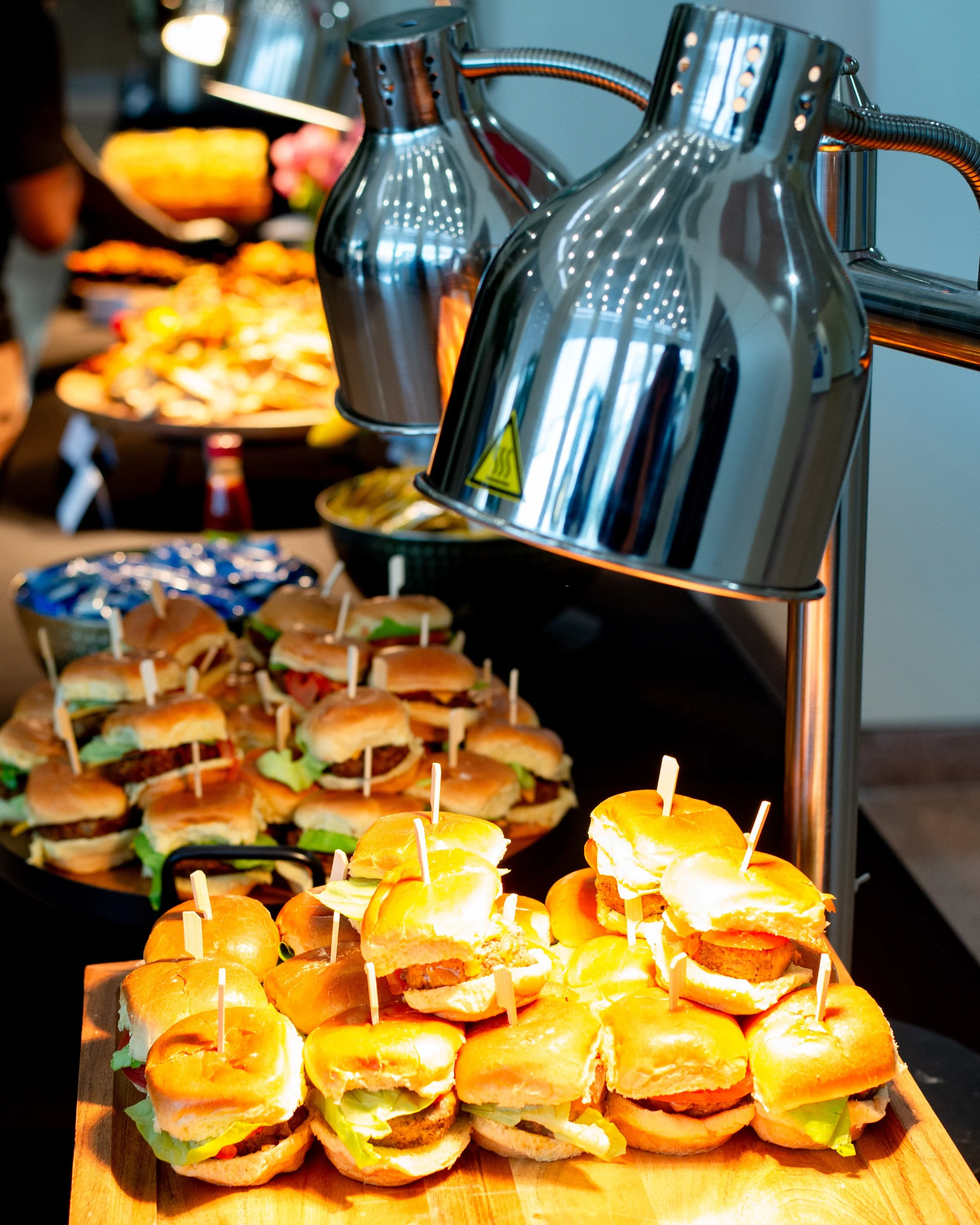 Assorted small sandwiches and sliders arranged on a table with metal heat lamps overhead at a buffet or catering event.