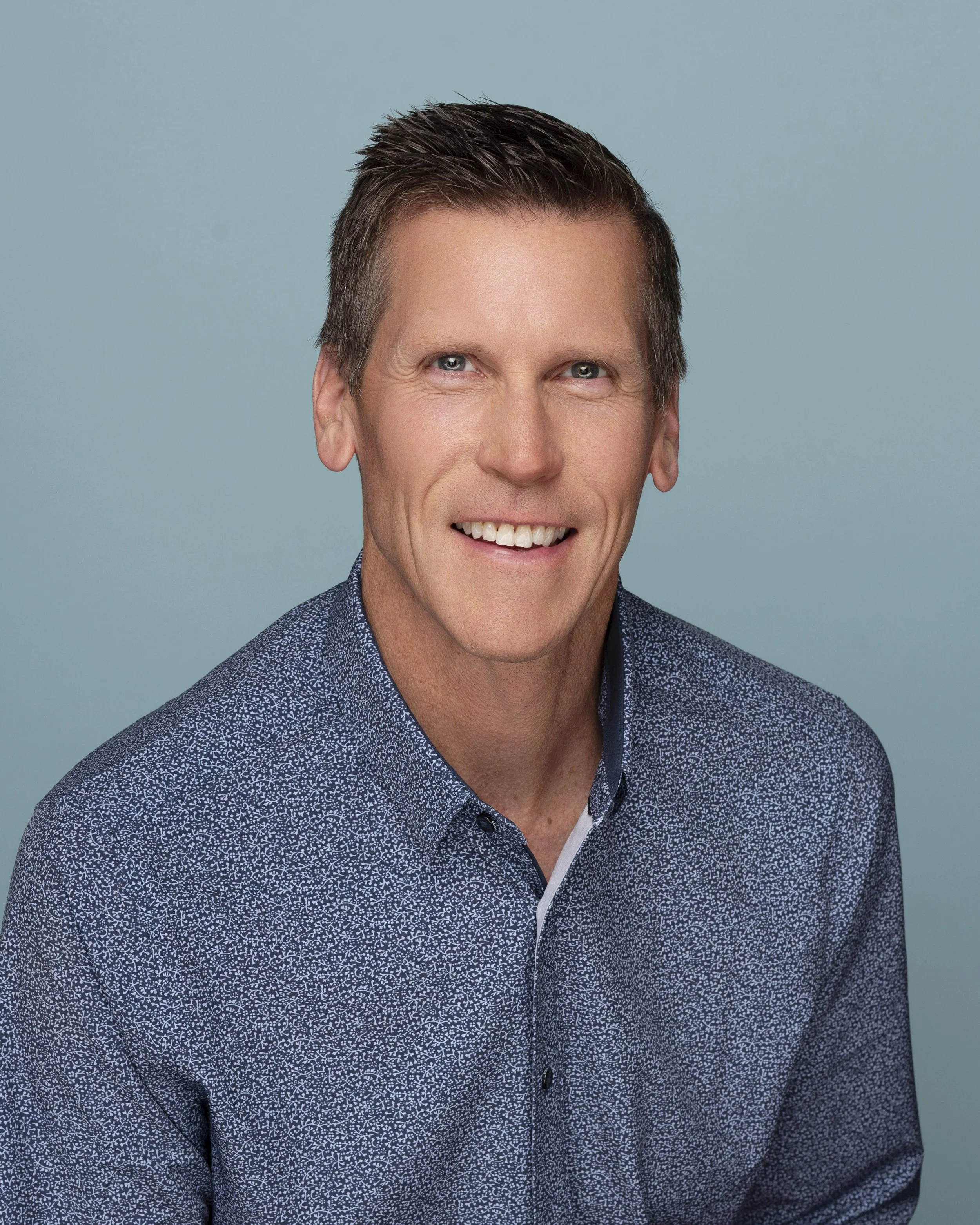 A smiling man with short brown hair, wearing a blue patterned collared shirt, against a light blue background.