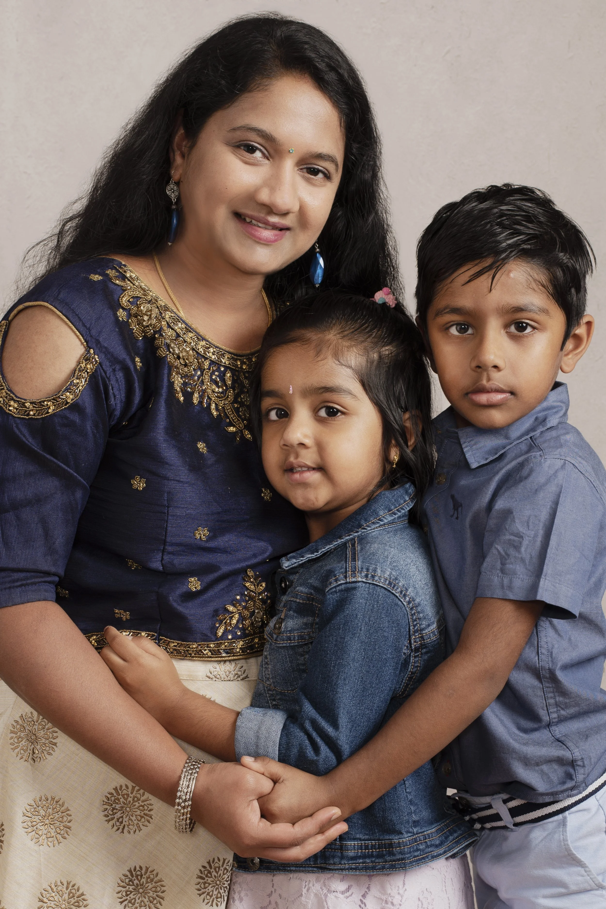 A woman with long black hair, wearing a blue embroidered top, is holding a young girl with black hair tied back, wearing a denim jacket. A young boy with short black hair, wearing a blue shirt, is standing beside them. They are all looking at the cam