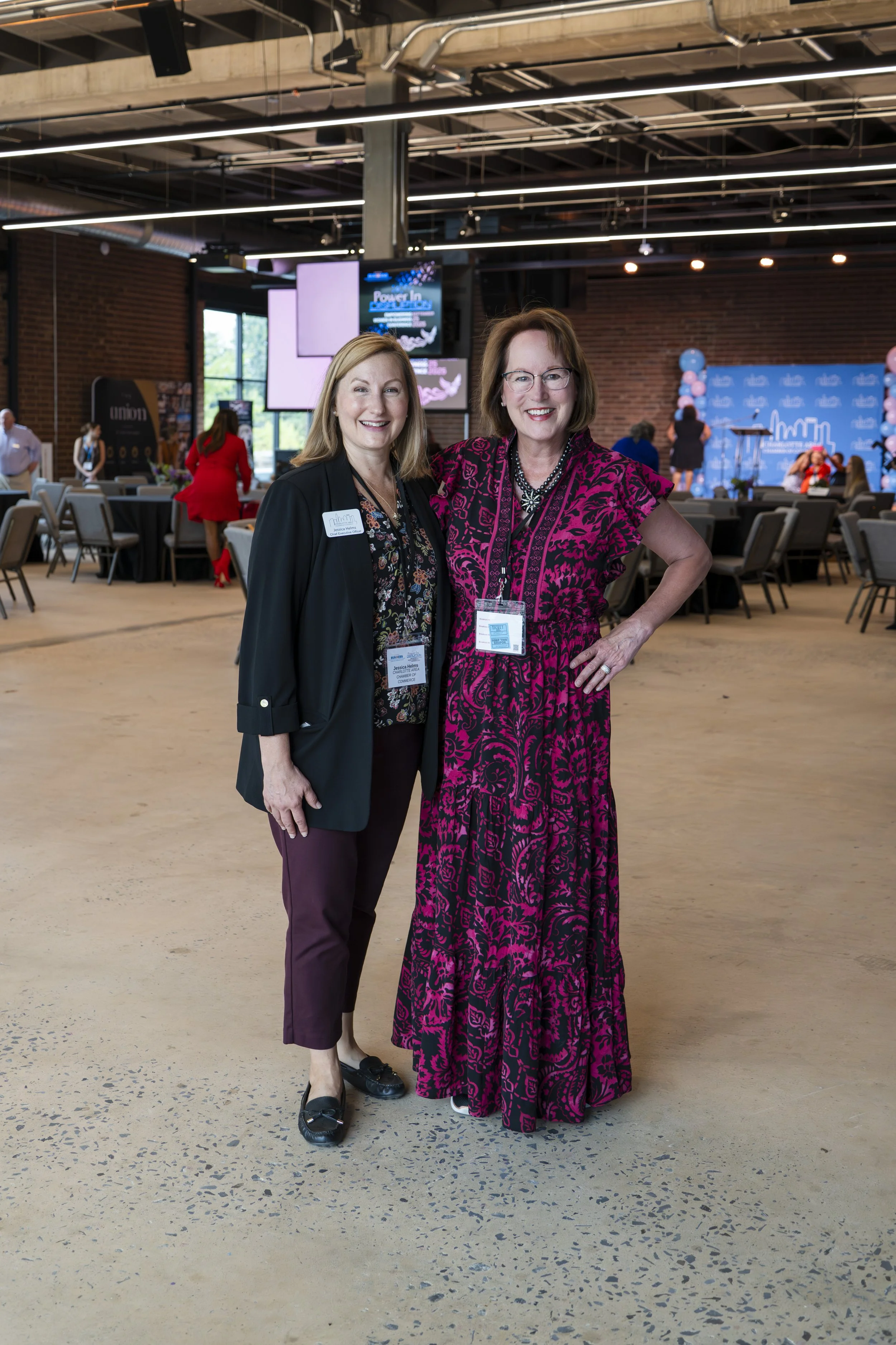 Two women standing together at an indoor event, smiling at the camera with a backdrop of an auditorium or conference hall with chairs, stage, and screens.