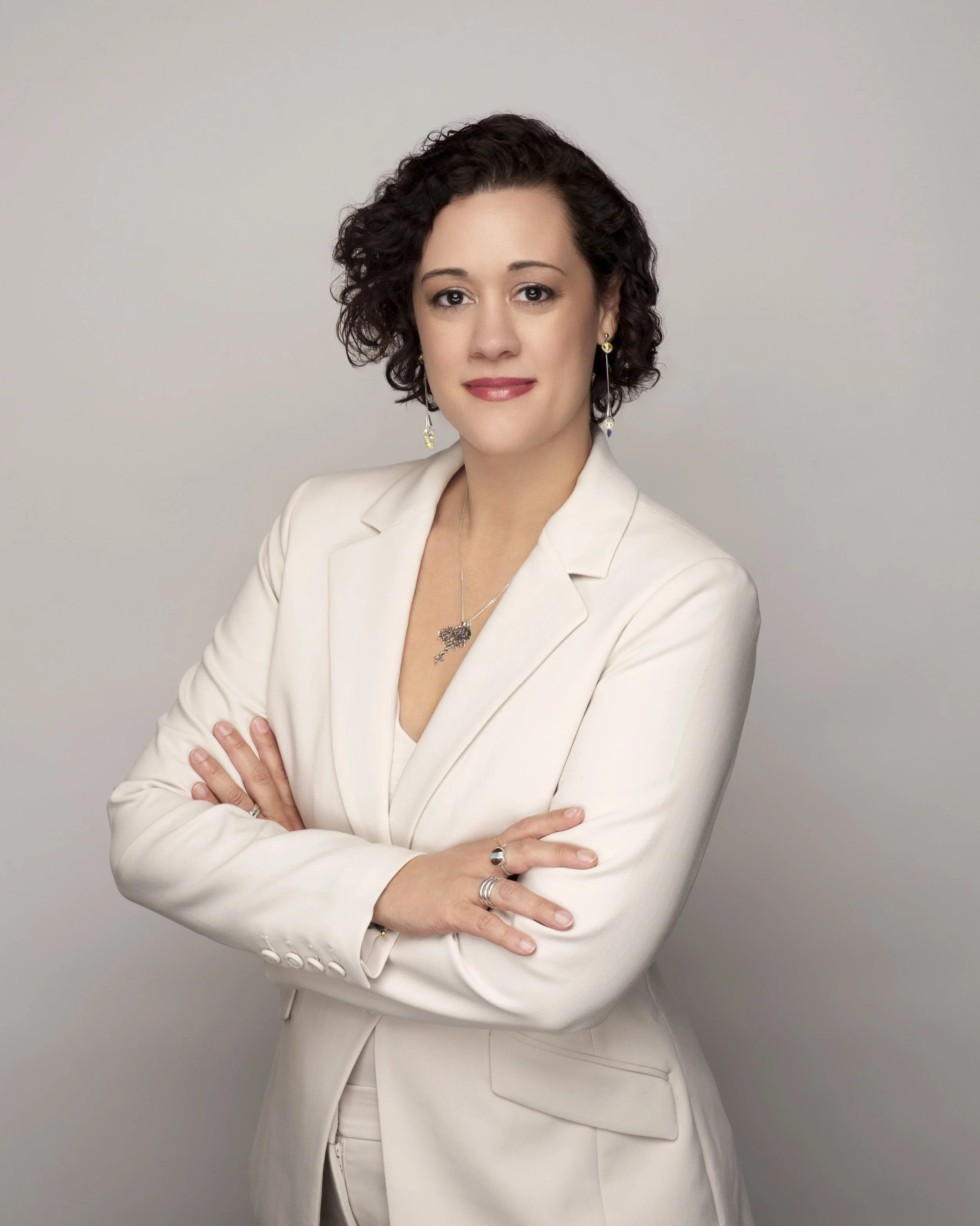A woman with short, curly dark hair and light makeup, wearing a cream-colored blazer and earrings, poses with arms crossed against a plain light gray background.