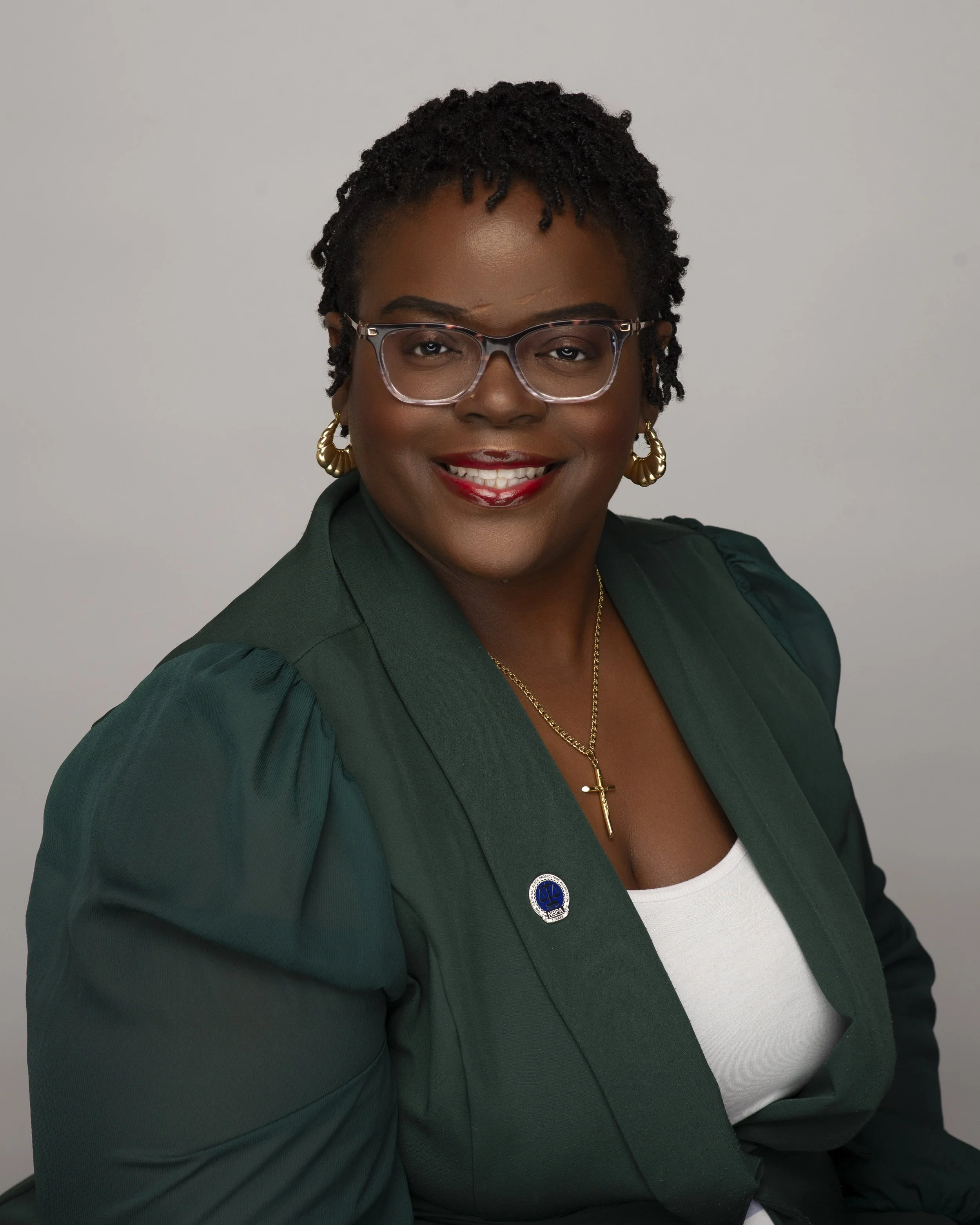 A professional woman with short, curly hair, wearing glasses, gold hoop earrings, a gold cross necklace, a green blazer with a pin, and a white top, smiling at the camera.