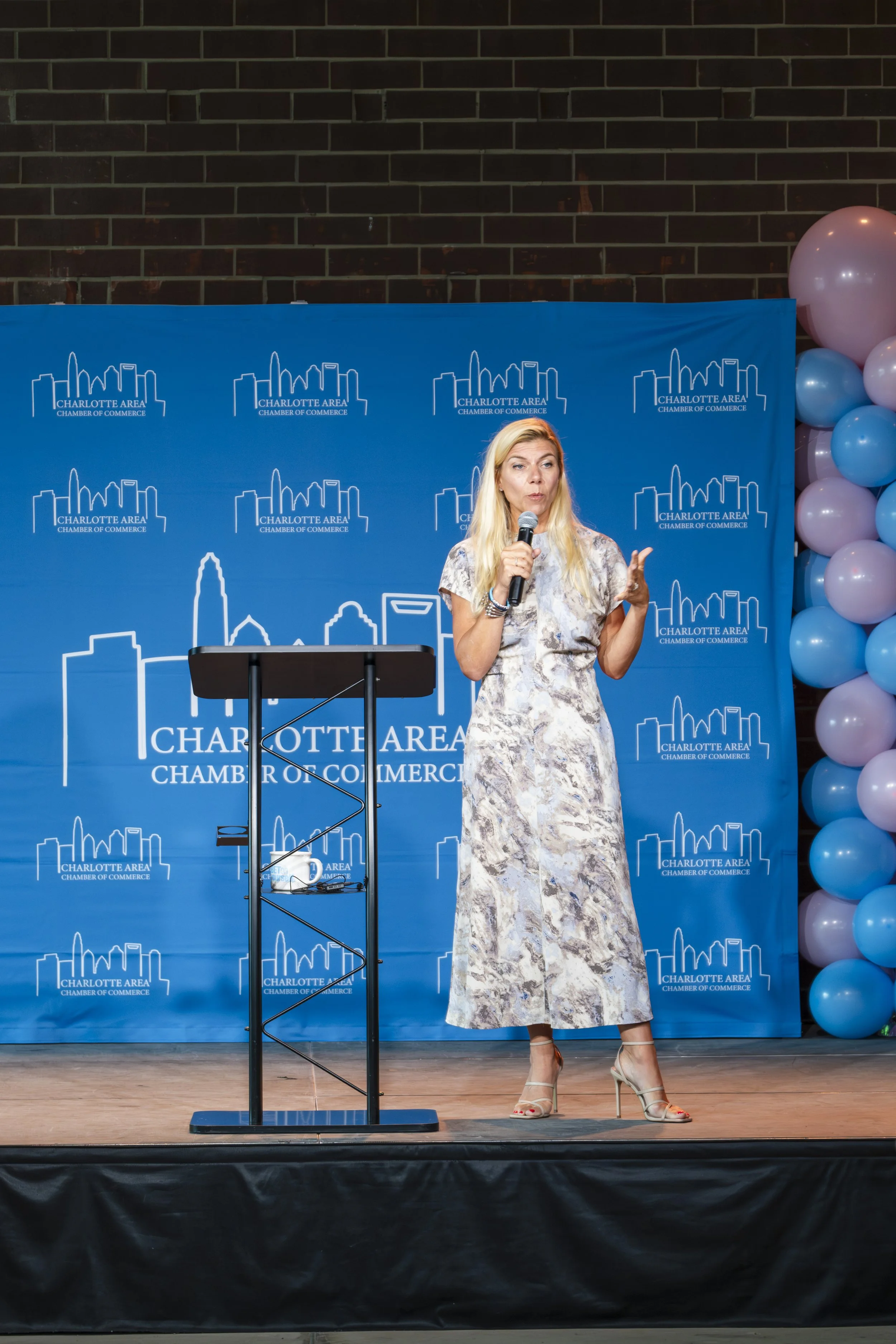 A woman speaking into a microphone on a stage with a blue Charlotte Area Chamber of Commerce backdrop and a cluster of purple and blue balloons on the right.