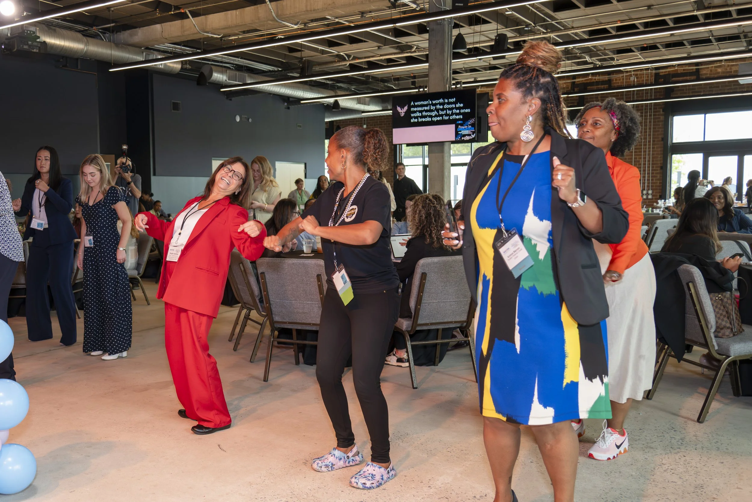 Several women are dancing and socializing at an indoor conference or event. They are wearing business casual and colorful outfits, with some wearing name badges. The background shows seated attendees, a large screen, and a modern, industrial-style sp