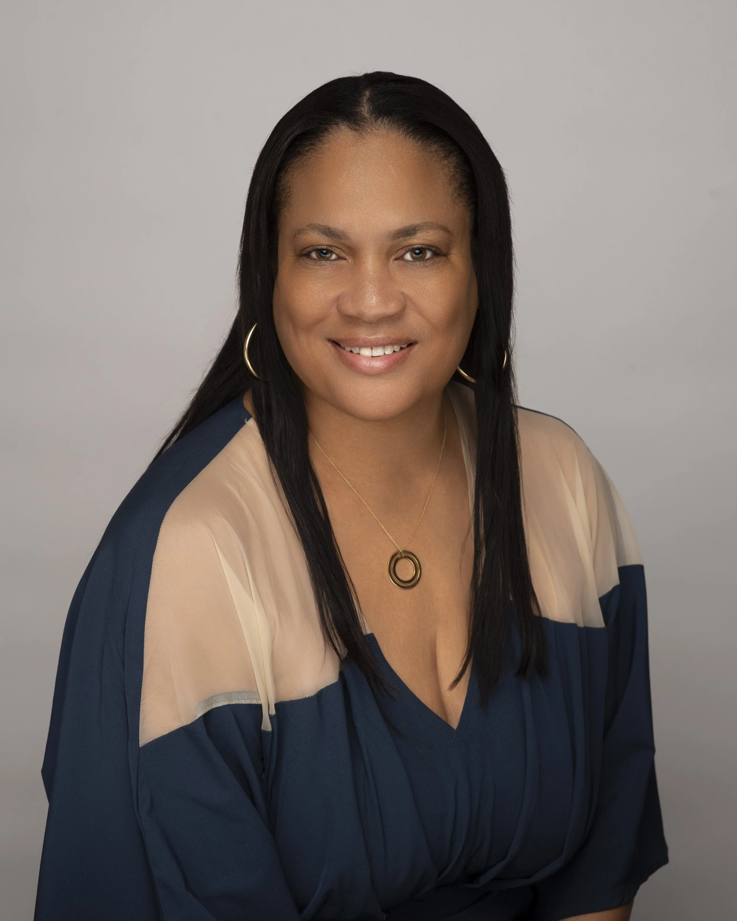 Portrait of a woman with medium skin tone, black hair, wearing hoop earrings, a necklace, in a navy and beige blouse, smiling against a plain gray background.