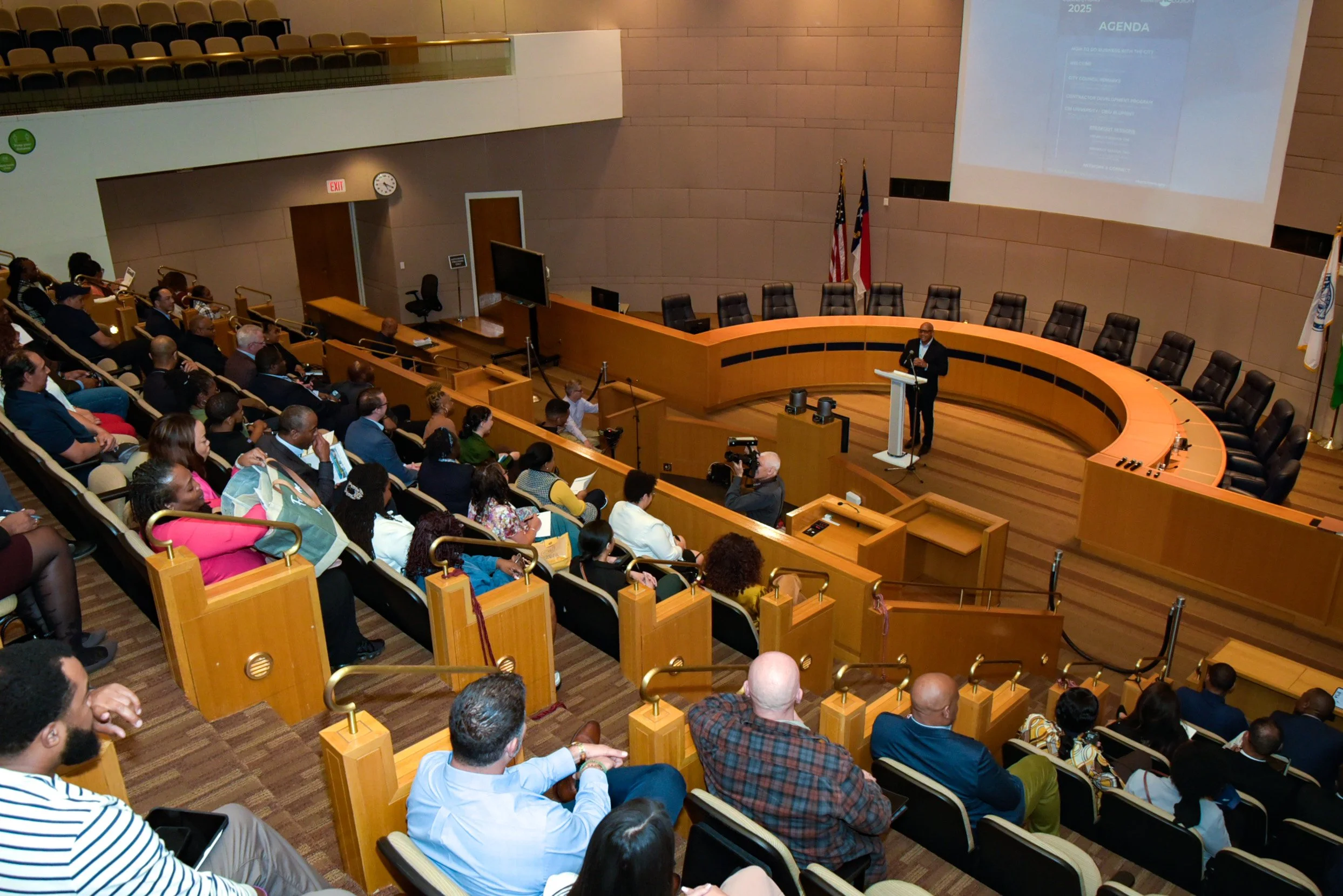 A formal presentation or conference is taking place in a large auditorium with a speaker standing at a podium on a circular stage. The audience is seated in tiered rows, and some are taking notes or recording the event. The room has a large projectio