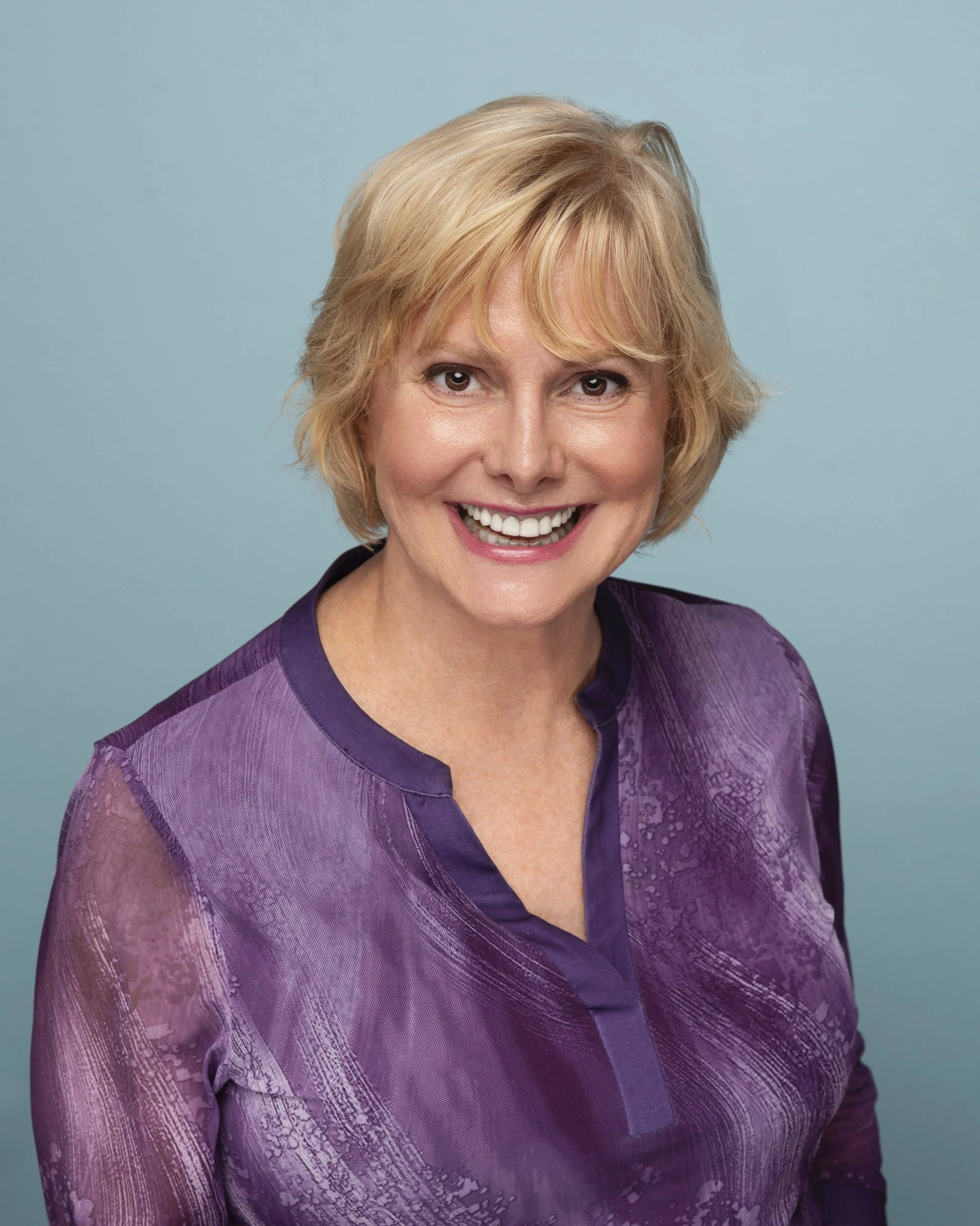 A smiling woman with short blonde hair wearing a purple blouse against a light blue background.