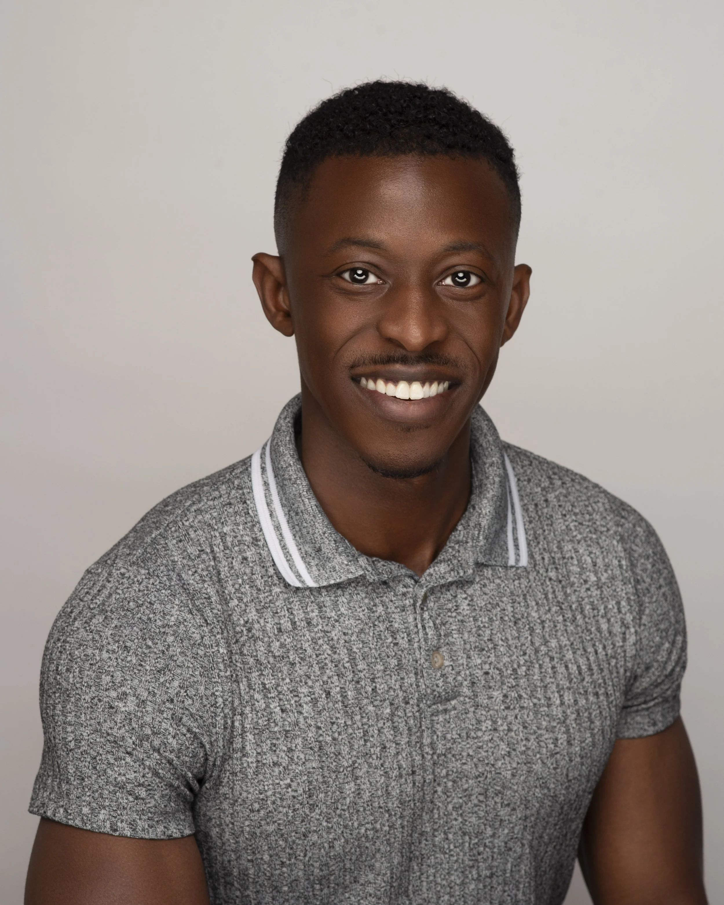 A young man with dark skin and short curly hair smiling, wearing a gray polo shirt with white stripes on the collar, against a plain gray background.