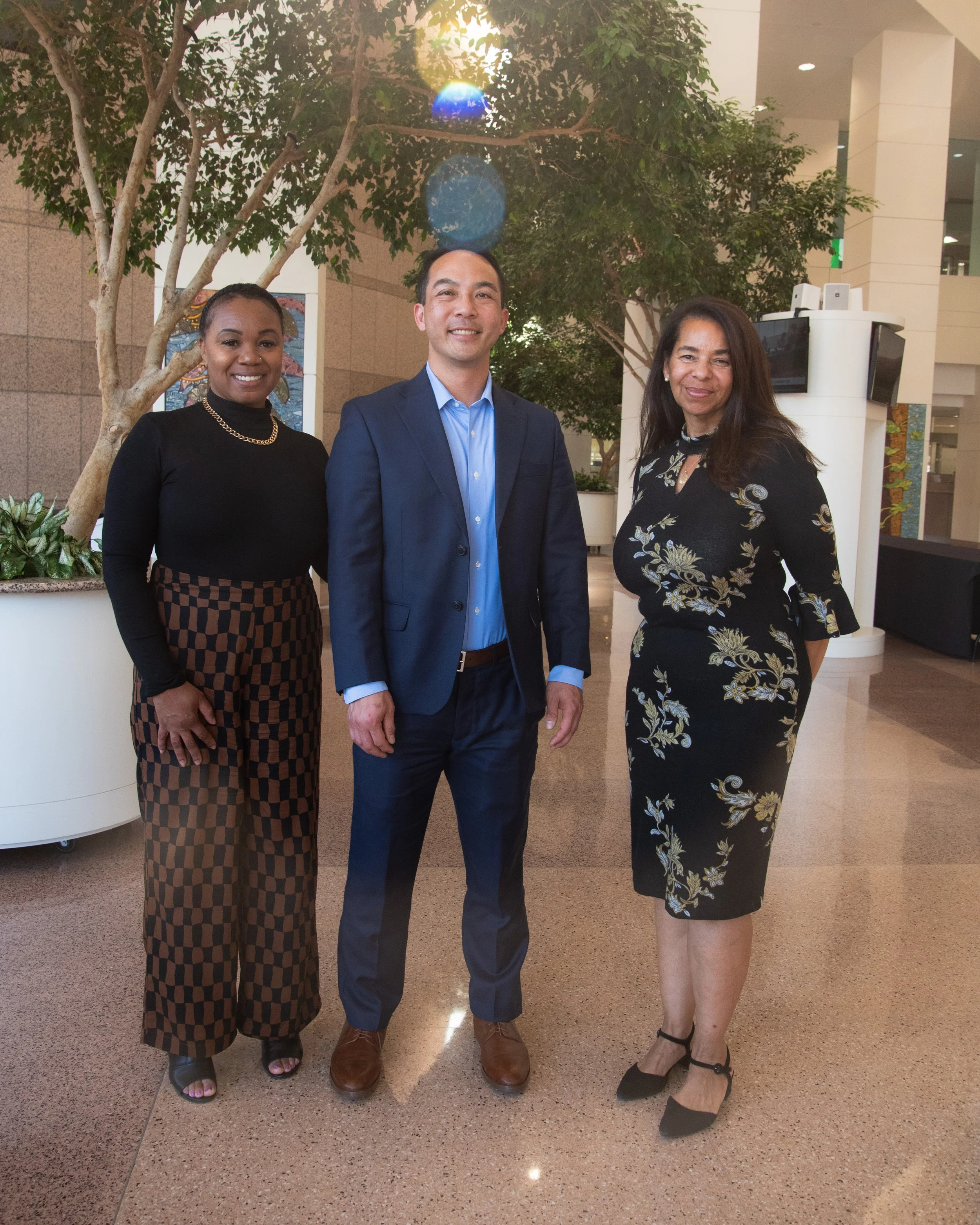 Three professionally dressed individuals standing in an indoor mall, smiling for a photo, with trees and a tiled floor in the background.