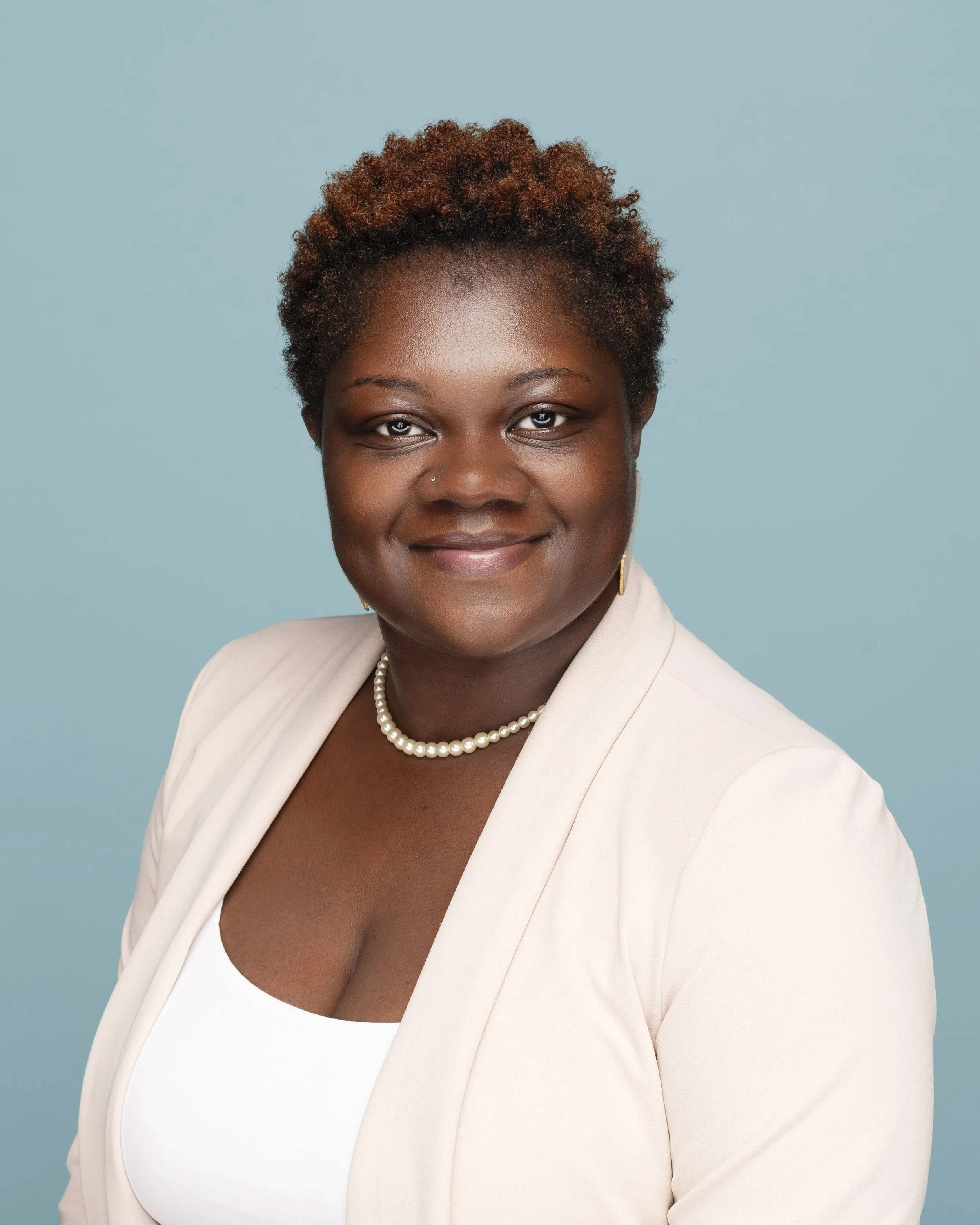Professional portrait of an African American woman with short curly hair, wearing a white top, a cream blazer, pearl necklace, and small earrings, standing against a light blue background.