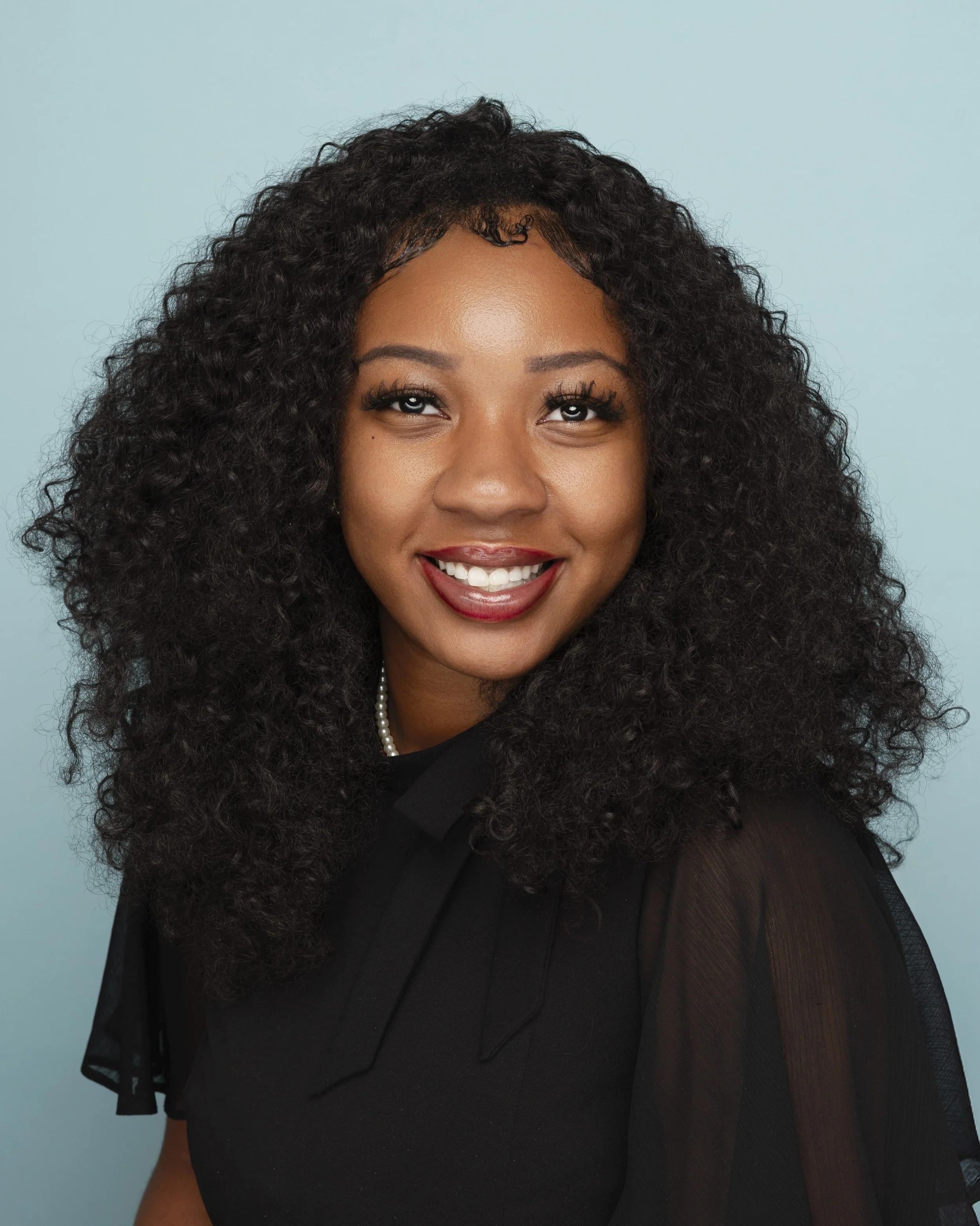 A young woman with dark curly hair smiling against a light blue background.