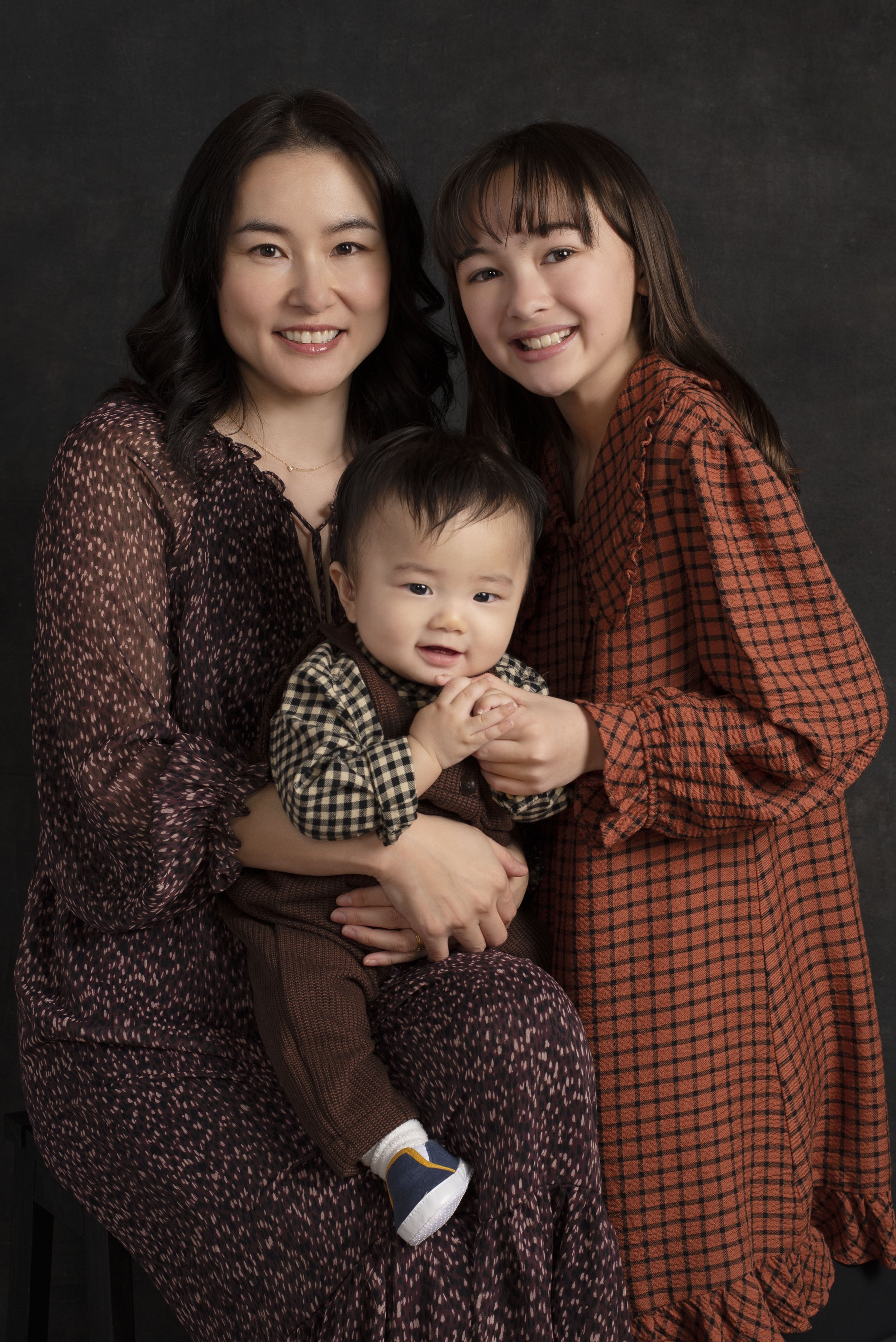 Three Asian females, two adults and one child, smiling and posing together against a dark background. The woman on the left is holding a young boy on her lap, and the woman on the right is leaning towards them, all wearing patterned clothing.