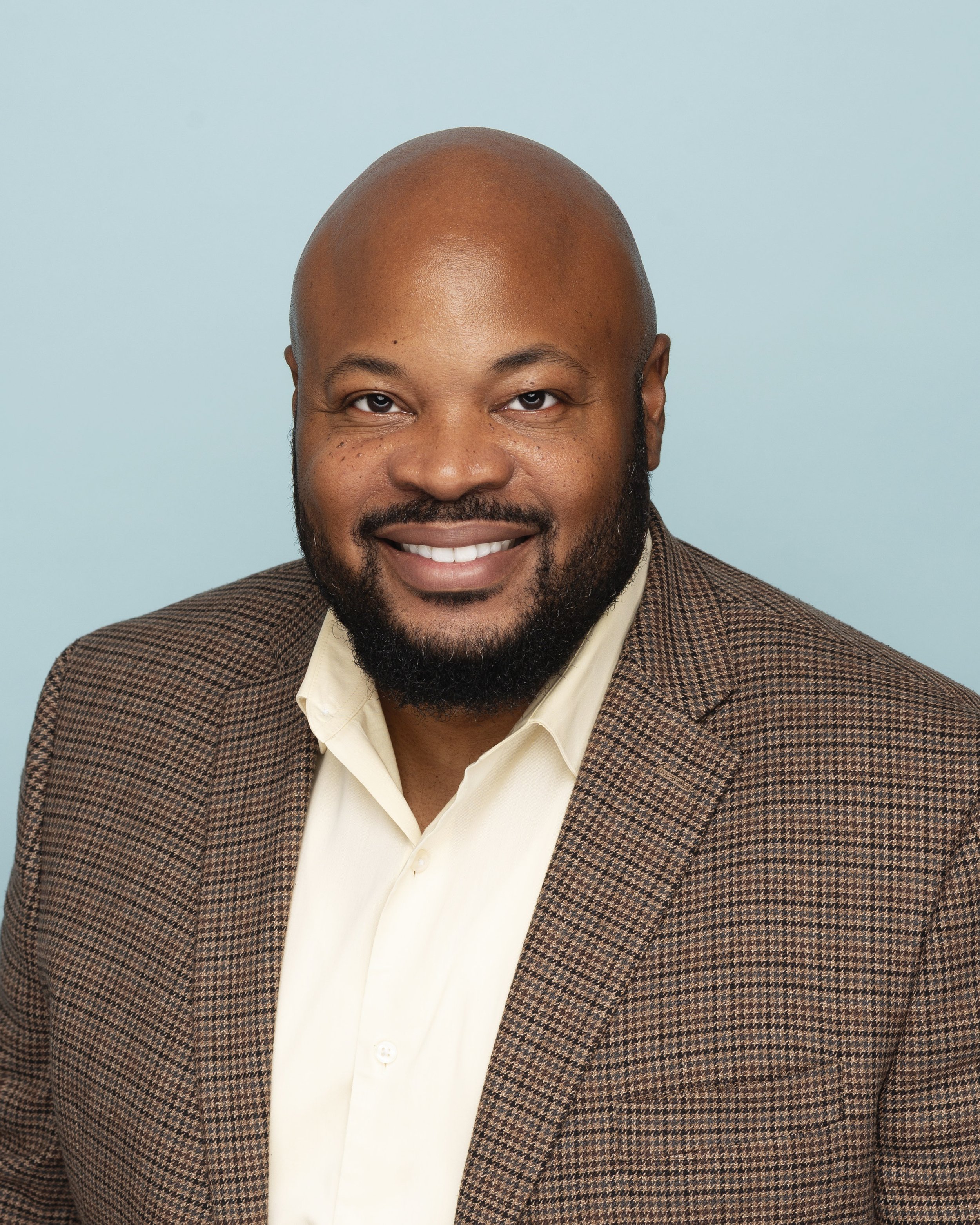 Portrait of a smiling African-American man with a bald head and a full beard, wearing a beige shirt and a brown checkered blazer, against a light blue background.