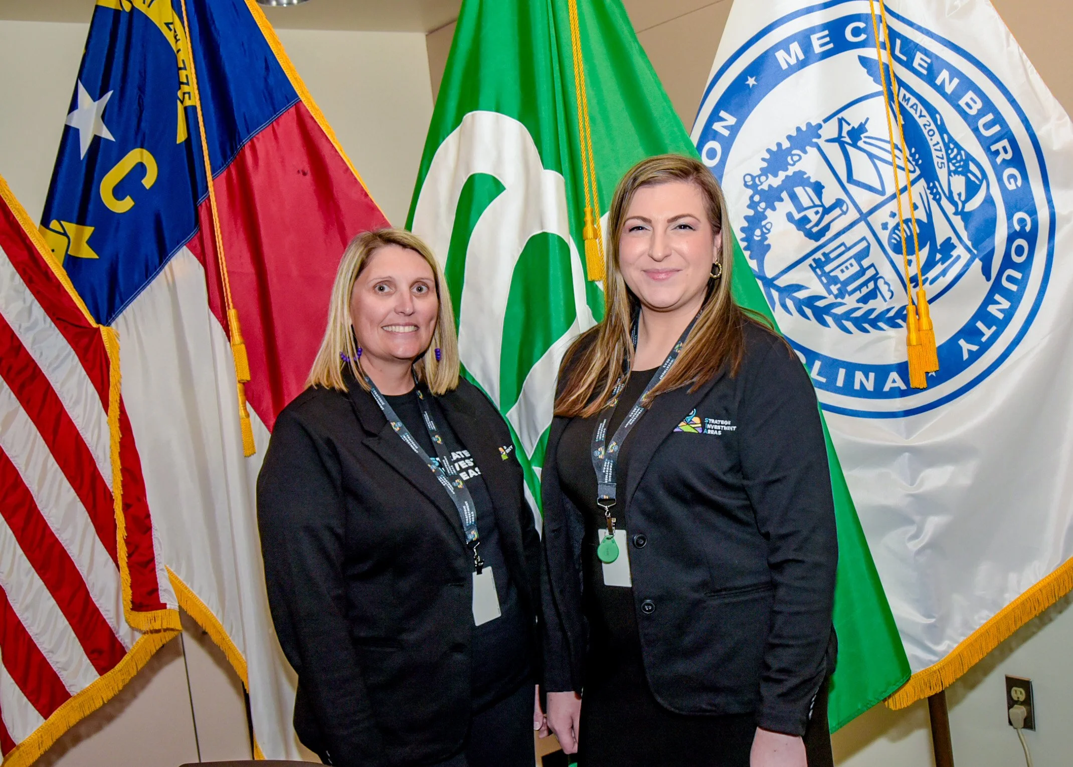 Two women standing in front of public service flags, including the U.S. flag, and official banners, smiling at the camera.