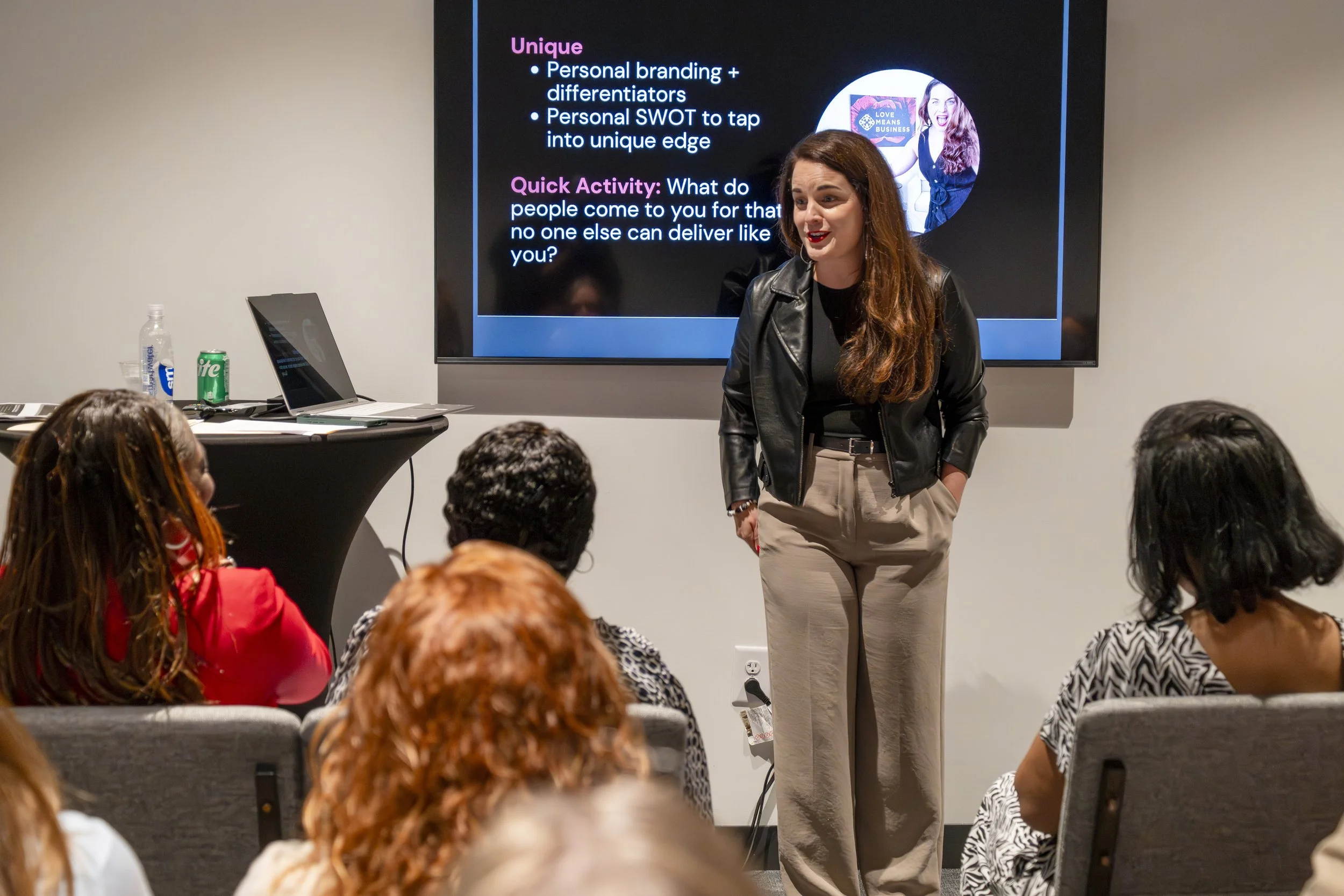 A woman in black leather jacket and beige pants giving a presentation to an audience, with a black screen behind her displaying text and images.