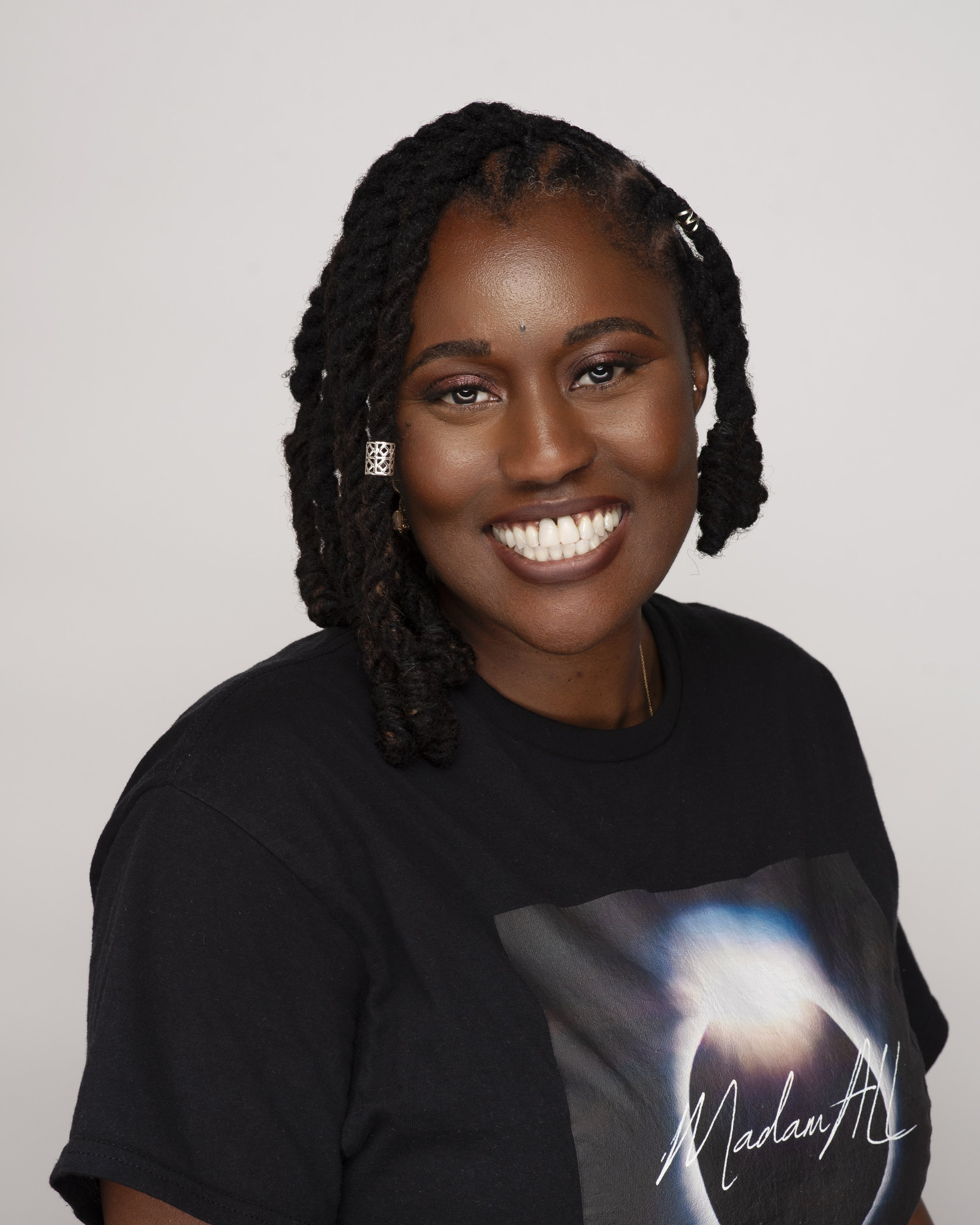 Smiling Black woman with short, twisted hair, wearing earrings, a necklace, and a black T-shirt with a galaxy design and the word "Madam" written on it, against a plain background.