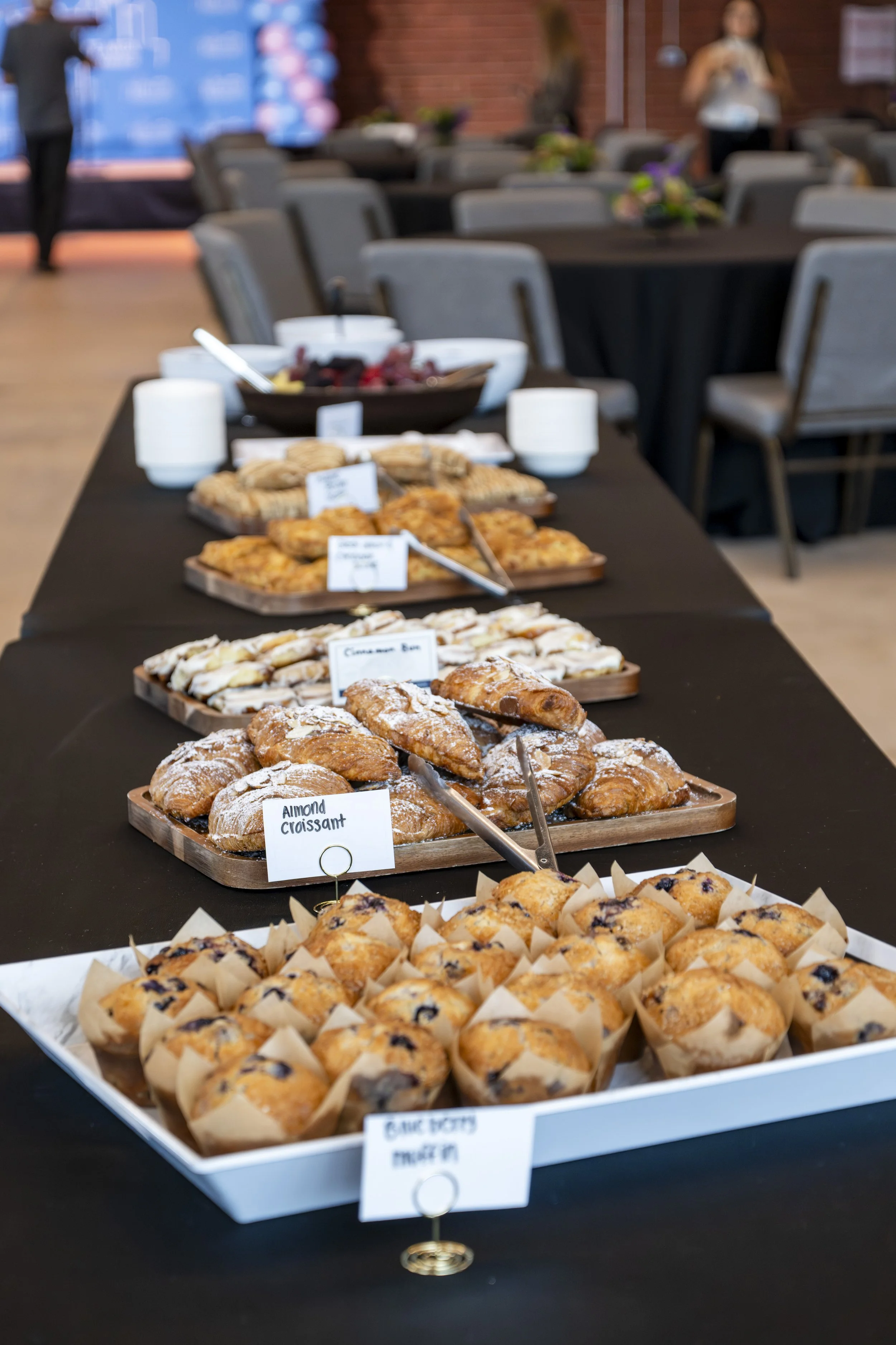 A table covered with a black cloth displaying assorted bakery items including muffins, croissants, cinnamon rolls, almond croissants, and berry muffins at a buffet or catering event, with blurred background of chairs and people.