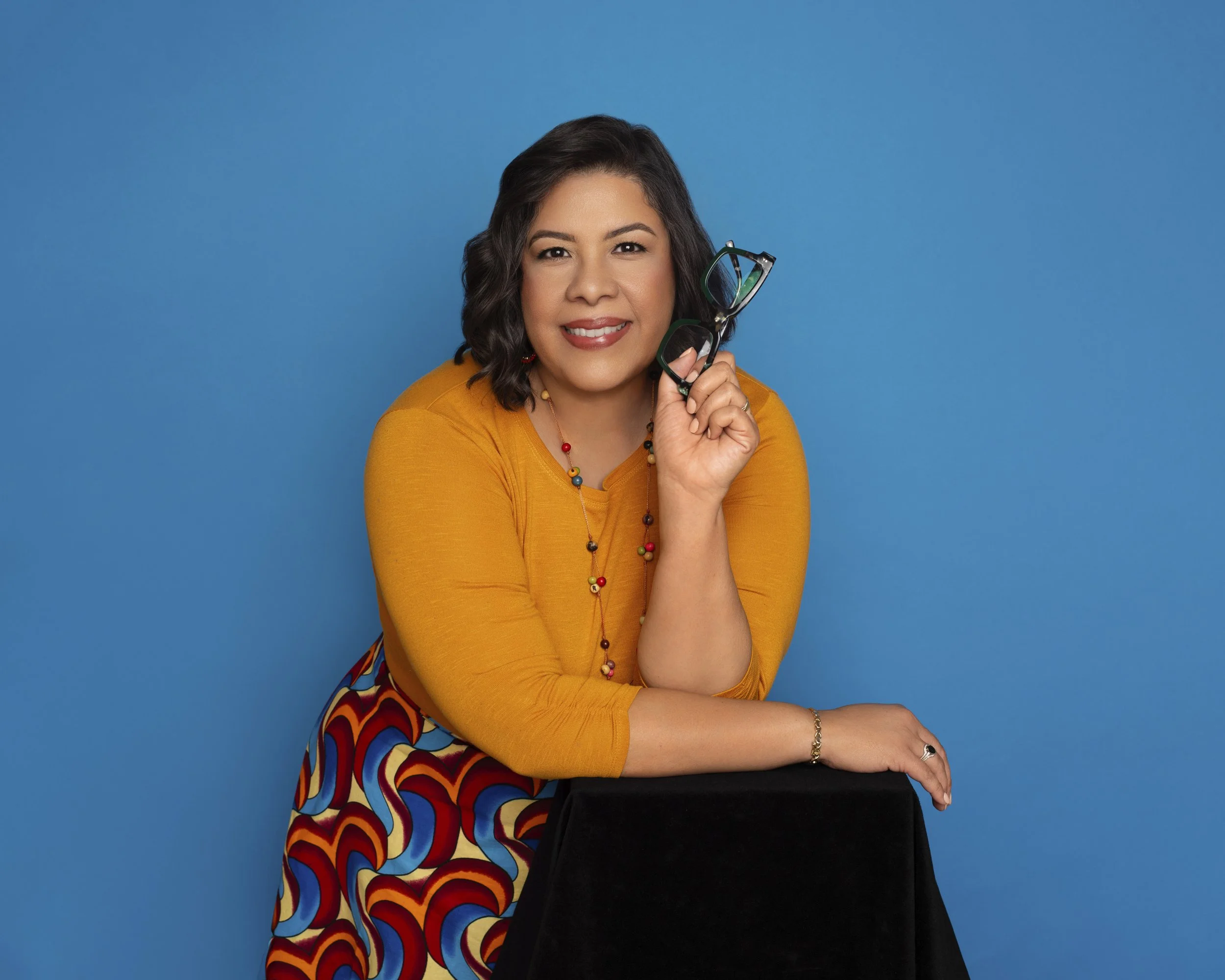 Woman in orange top holding glasses, smiling, with blue background.