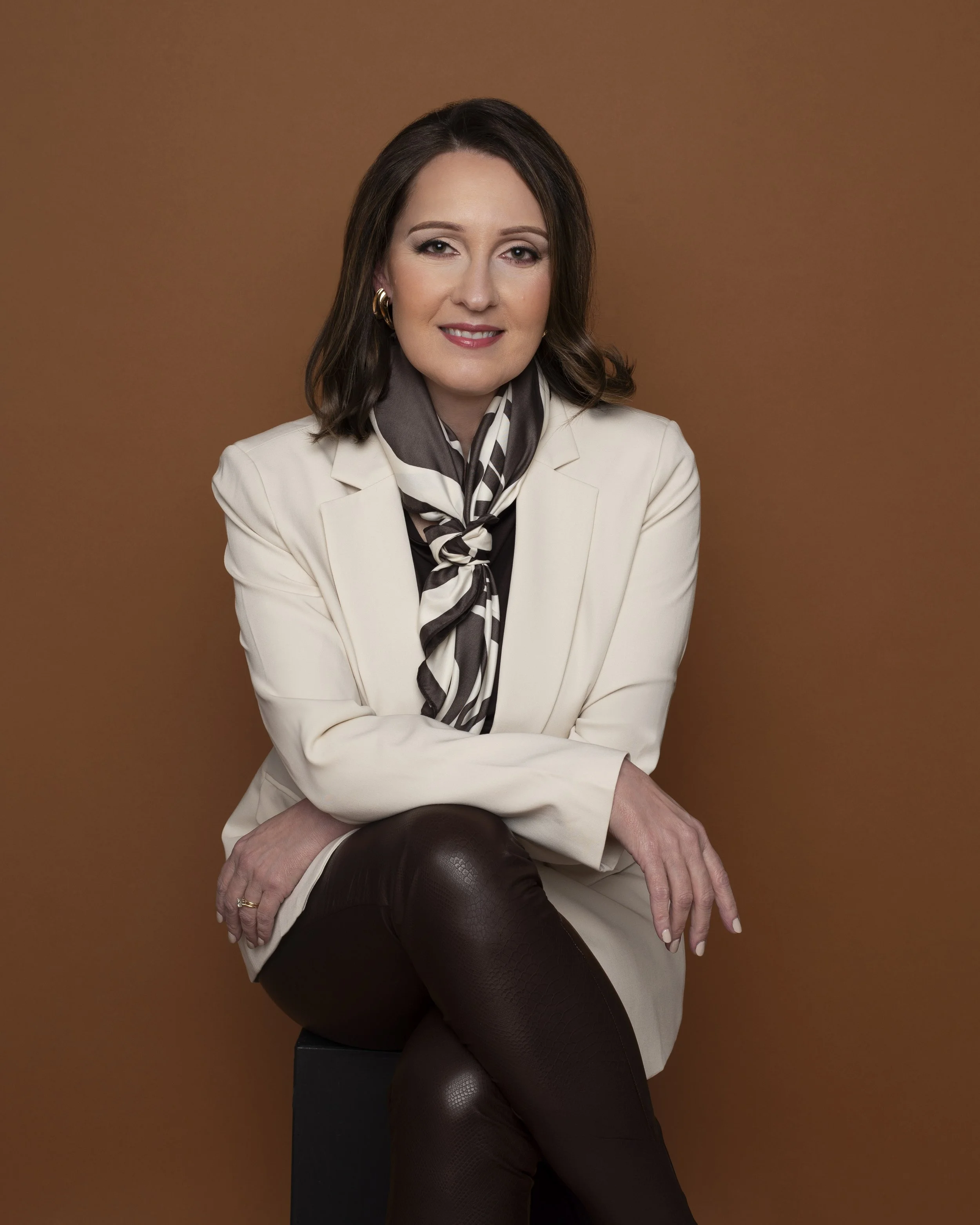 A woman with shoulder-length brown hair sitting on a black stool against a brown background, wearing a white blazer, black pants, and a patterned scarf.