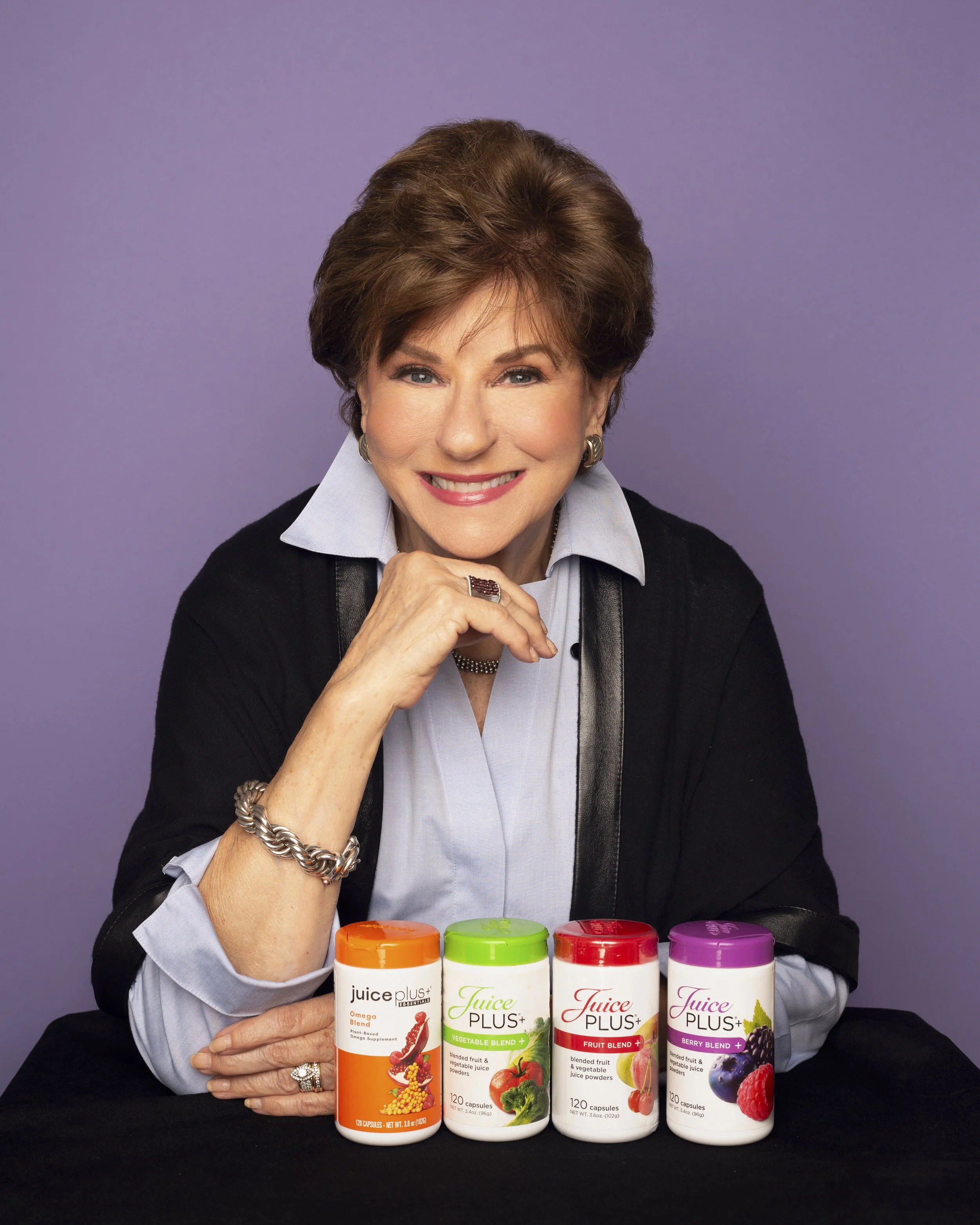 A woman smiling and sitting at a table with four bottles of Juice Plus supplements, purple background.