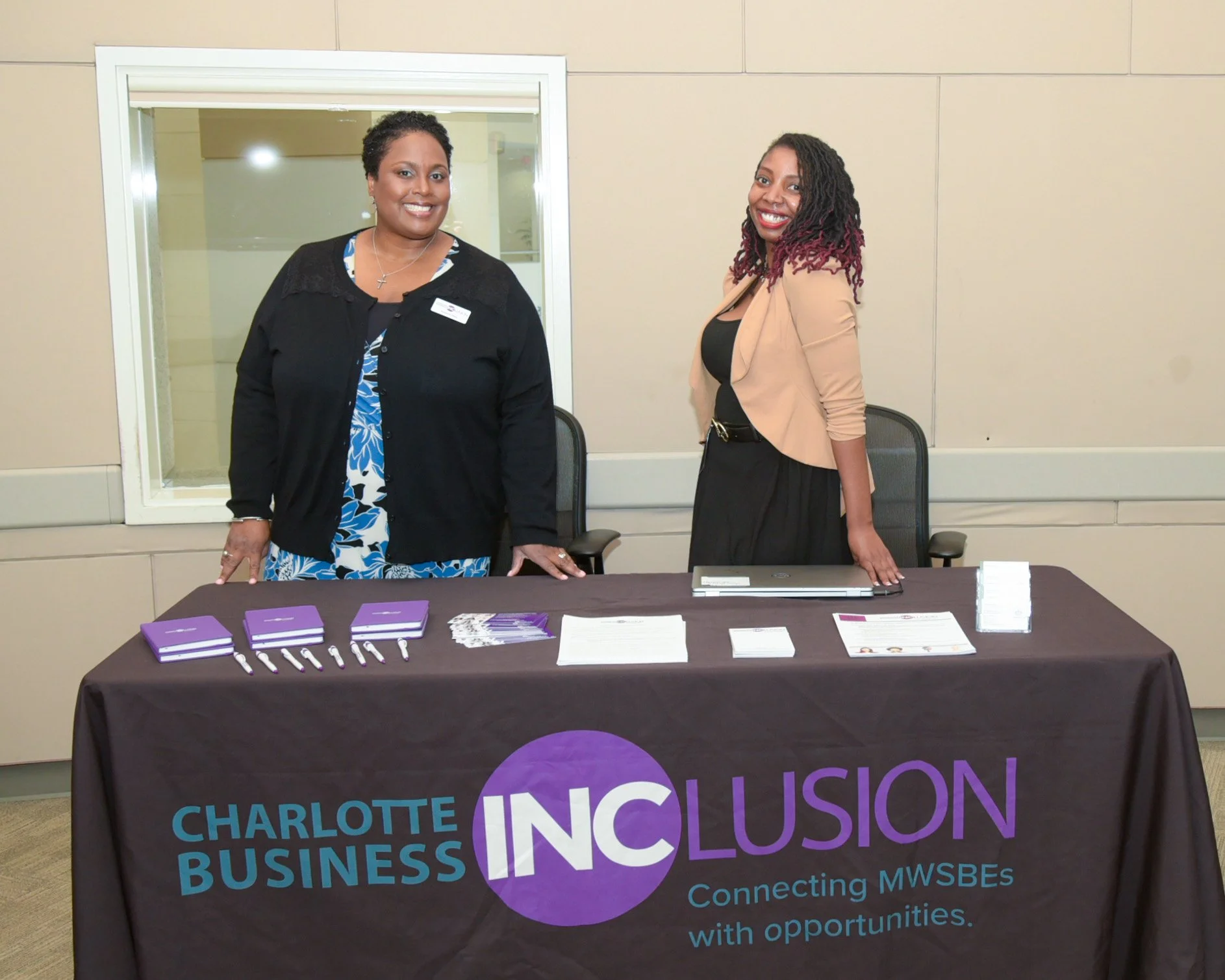 Two women standing behind a table with Charlotte Business INCLUSION branding, promotional materials, and pens, in an indoor setting.
