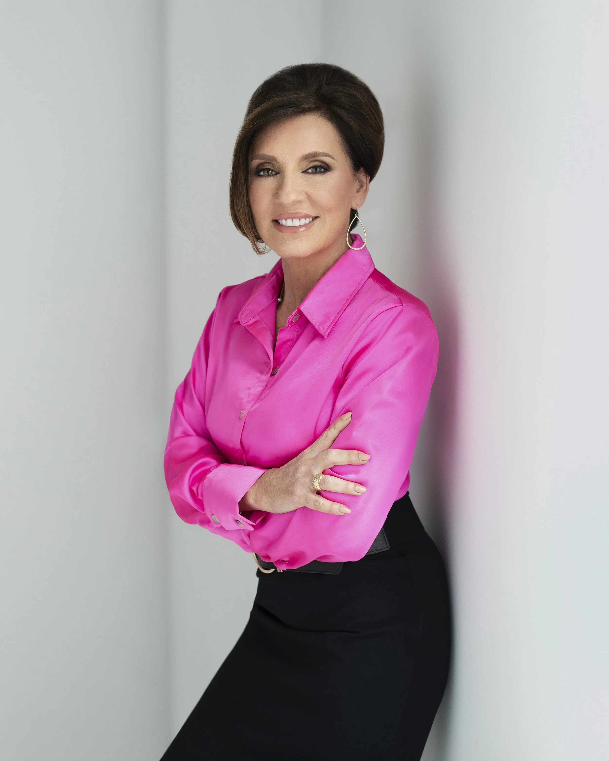 A woman with short brown hair wearing a bright pink blouse and black skirt, standing against a white wall with her arms crossed, smiling at the camera.