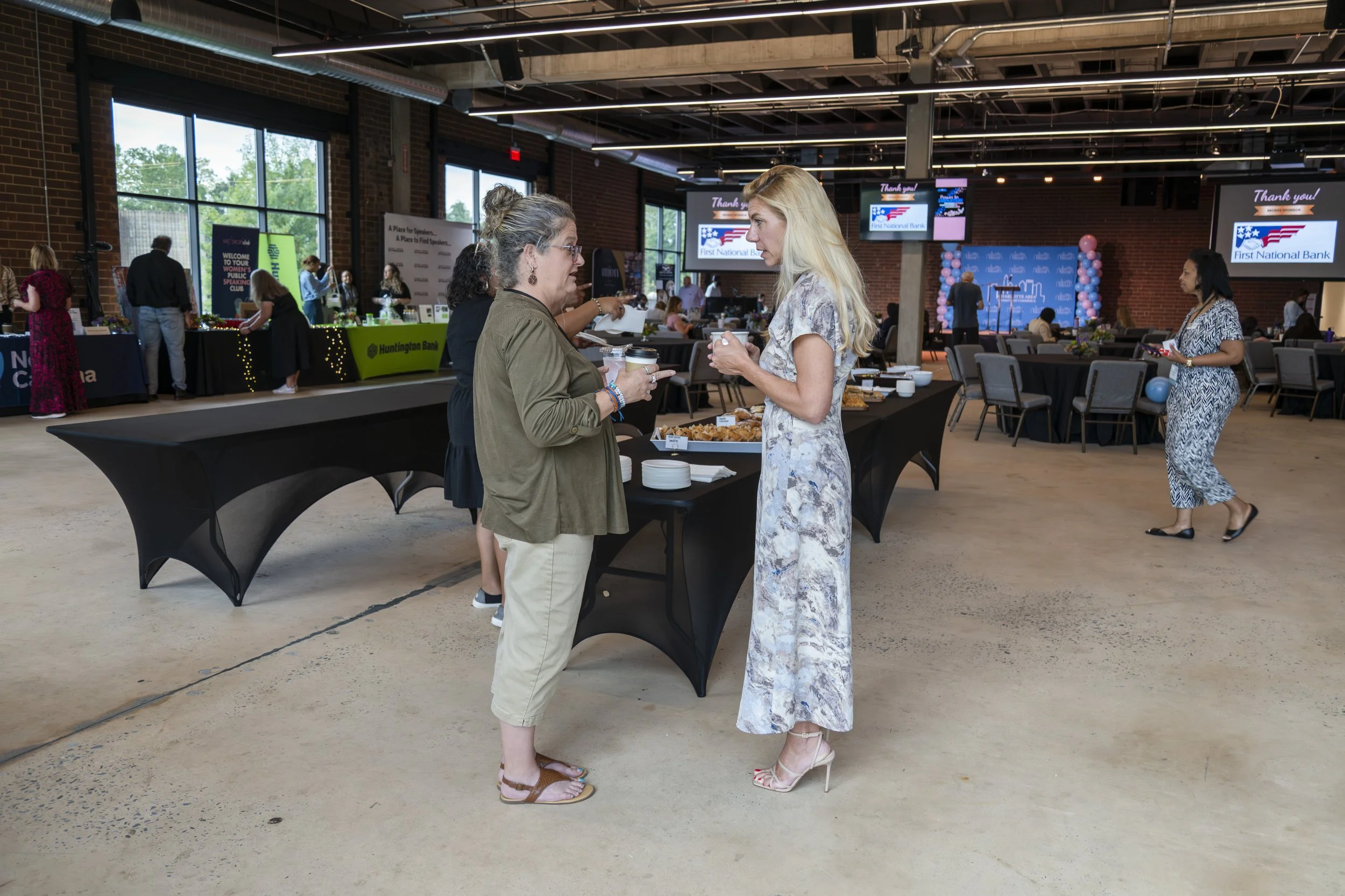 Women conversing at a buffet table during an event in a spacious venue with brick walls and large windows.