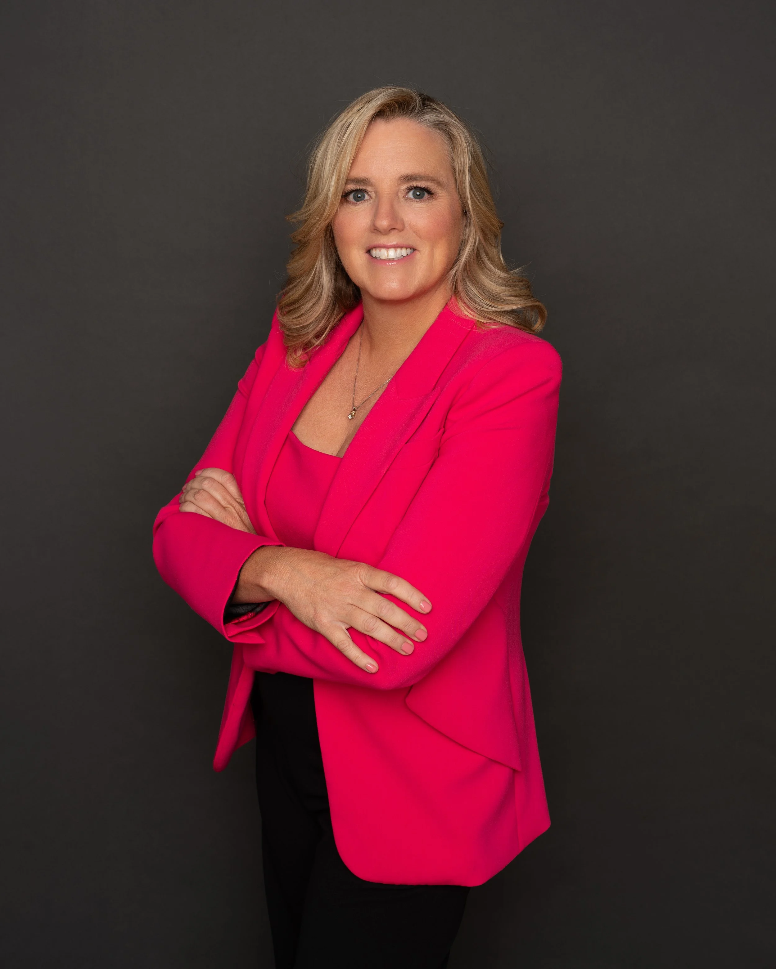 A woman with blonde, shoulder-length hair and blue eyes, smiling, dressed in a pink blazer and top, standing against a dark gray background.
