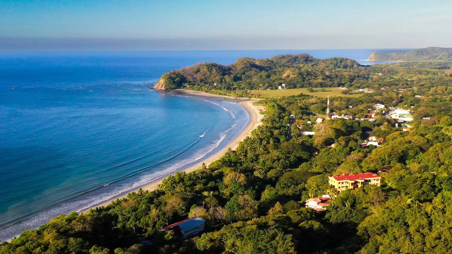 Aerial view of a tropical beach with a curved shoreline, turquoise water, and lush green trees lining the coast. Small buildings are visible among the trees, with hills and distant landforms in the background under a clear sky.