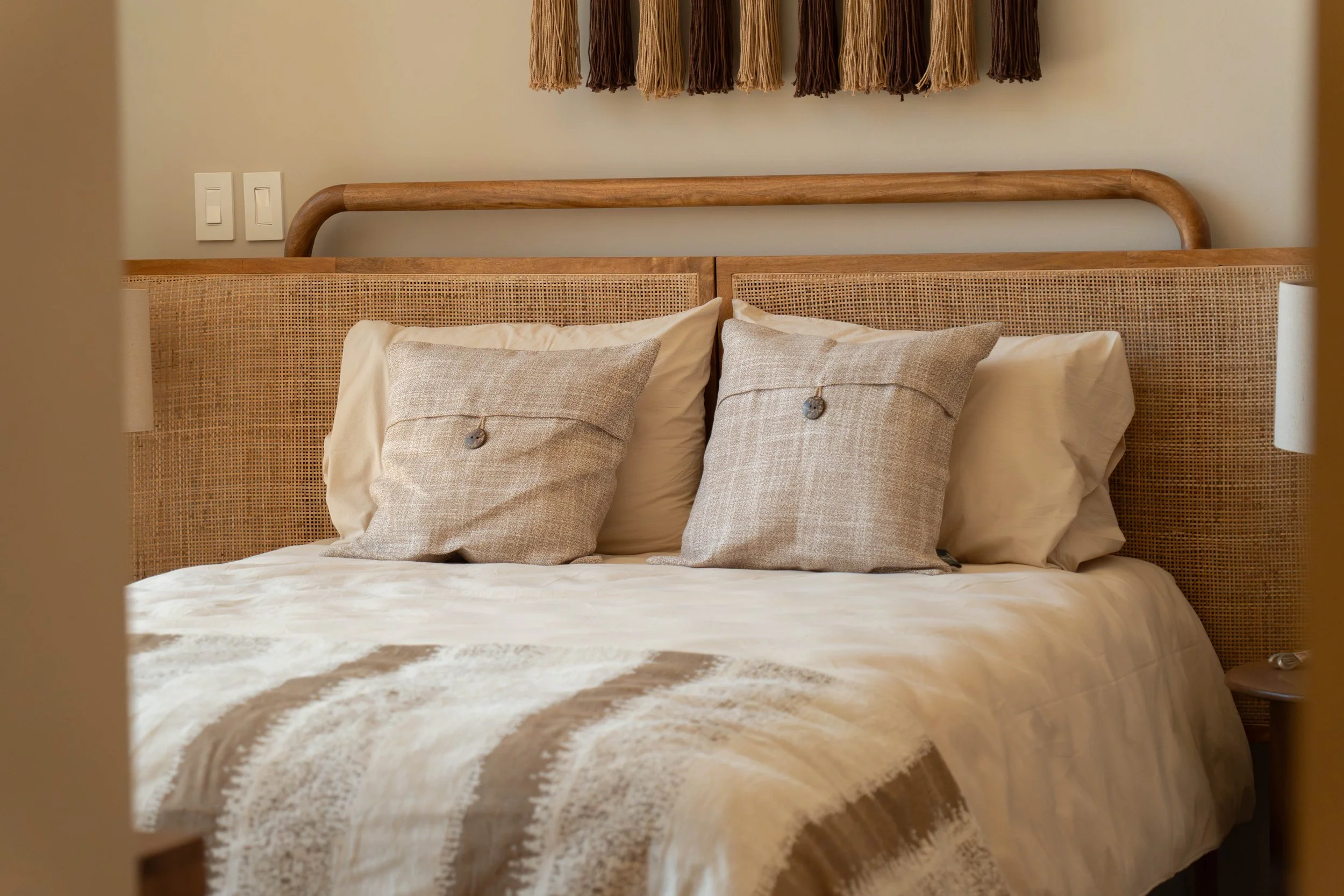 A neatly made bed with beige and cream pillows, a wooden headboard, and a textured beige and white blanket. A woven wall hanging with shades of brown, beige, and tan decorates the wall above the bed. Two white electrical switches are visible on the w